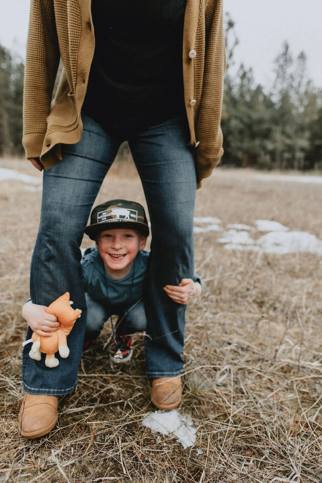 A person in a brown jacket, black shirt, and jeans standing outdoors with a child crouching between their legs. The child is smiling, wearing a cap, blue jacket, and holding a small plush toy. The ground is dry grass with patches of snow, and trees are blurred in the background.
