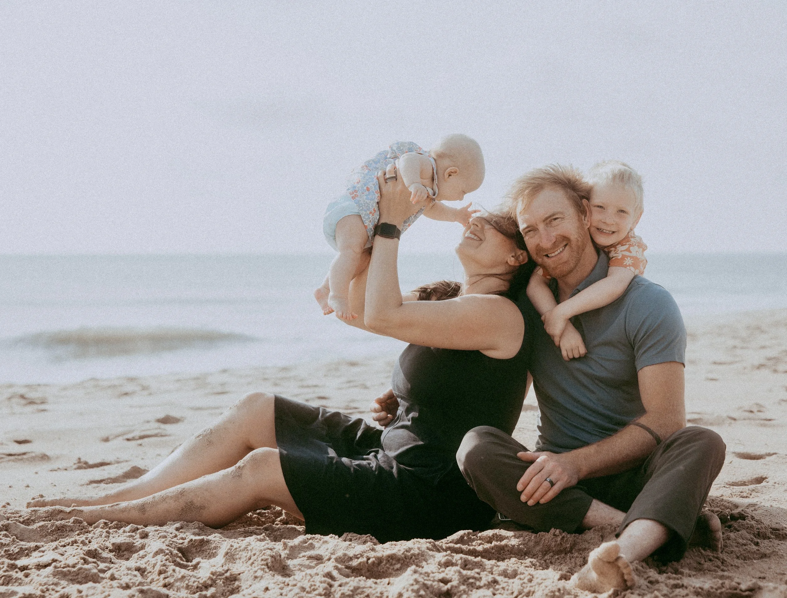 A family on the beach smiling and playing together. The mother and father are seated on the sand, with the mother holding a baby and the father embracing their older child.
