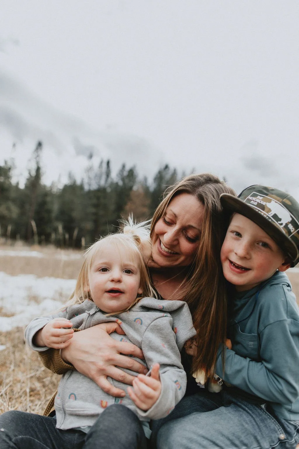 A healthy mom with two young children, a girl and a boy, outdoors in a field with snow and trees in the background, all smiling and enjoying the moment.