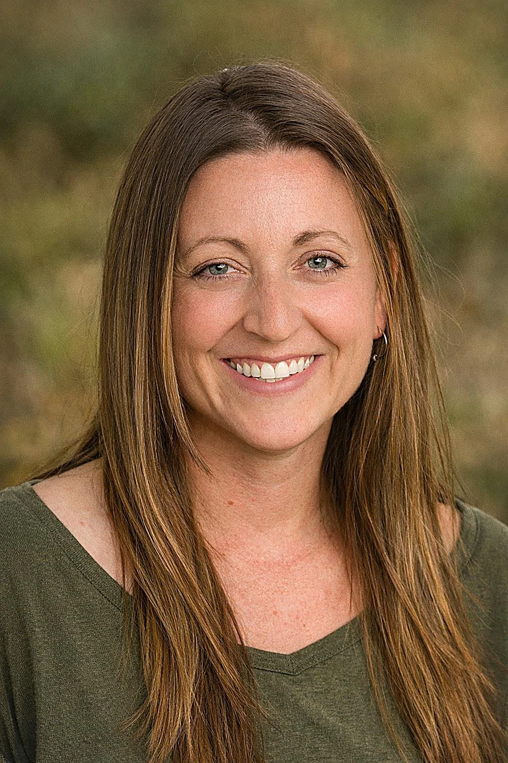 A woman with long brown hair, blue eyes, smiling, wearing an olive green top, outdoors with blurred greenery background.