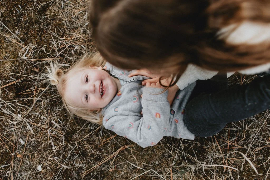 A healthy young girl with blonde hair lying on the ground, smiling and looking at the camera, while a woman with brown hair is leaning over her, holding her hands.