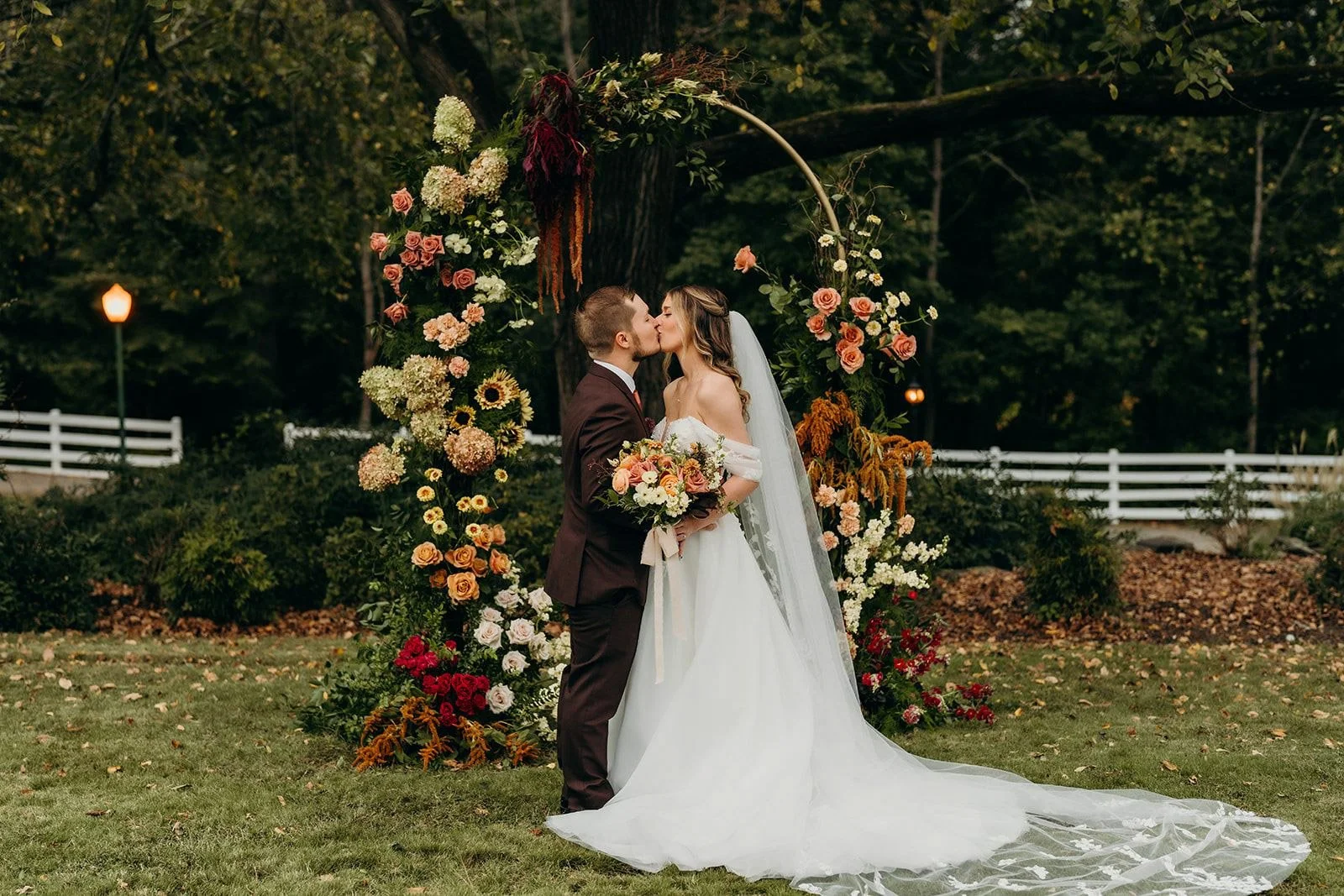Bride and groom kissing in front of a circular fall floral arch with hydrangea, roses, sunflowers, and hanging amaranthus at The Wakefield Barn near Raleigh