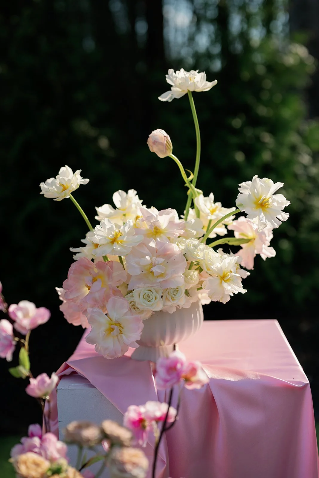 A floral arrangement of white and pale pink flowers in a white vase, placed on a pink tablecloth outdoors with dark, blurred background.