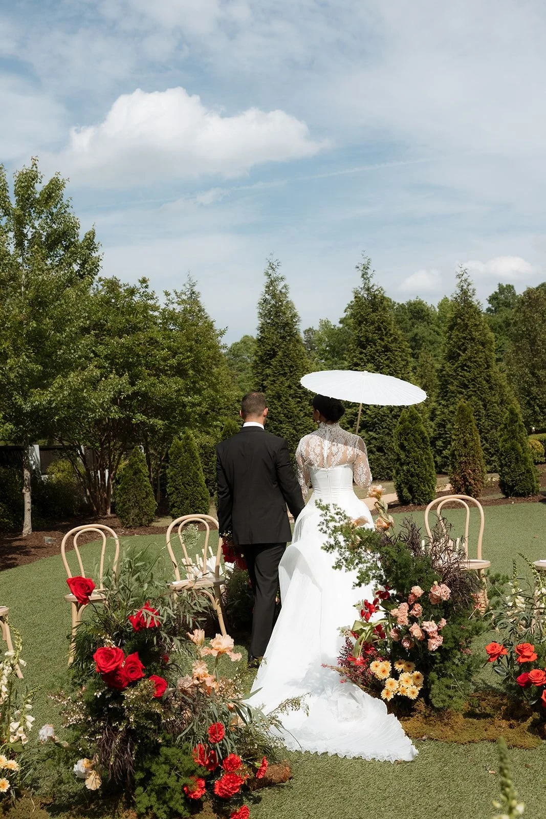 A bride and groom walking down an outdoor aisle decorated with flowers, with the bride holding a white parasol.