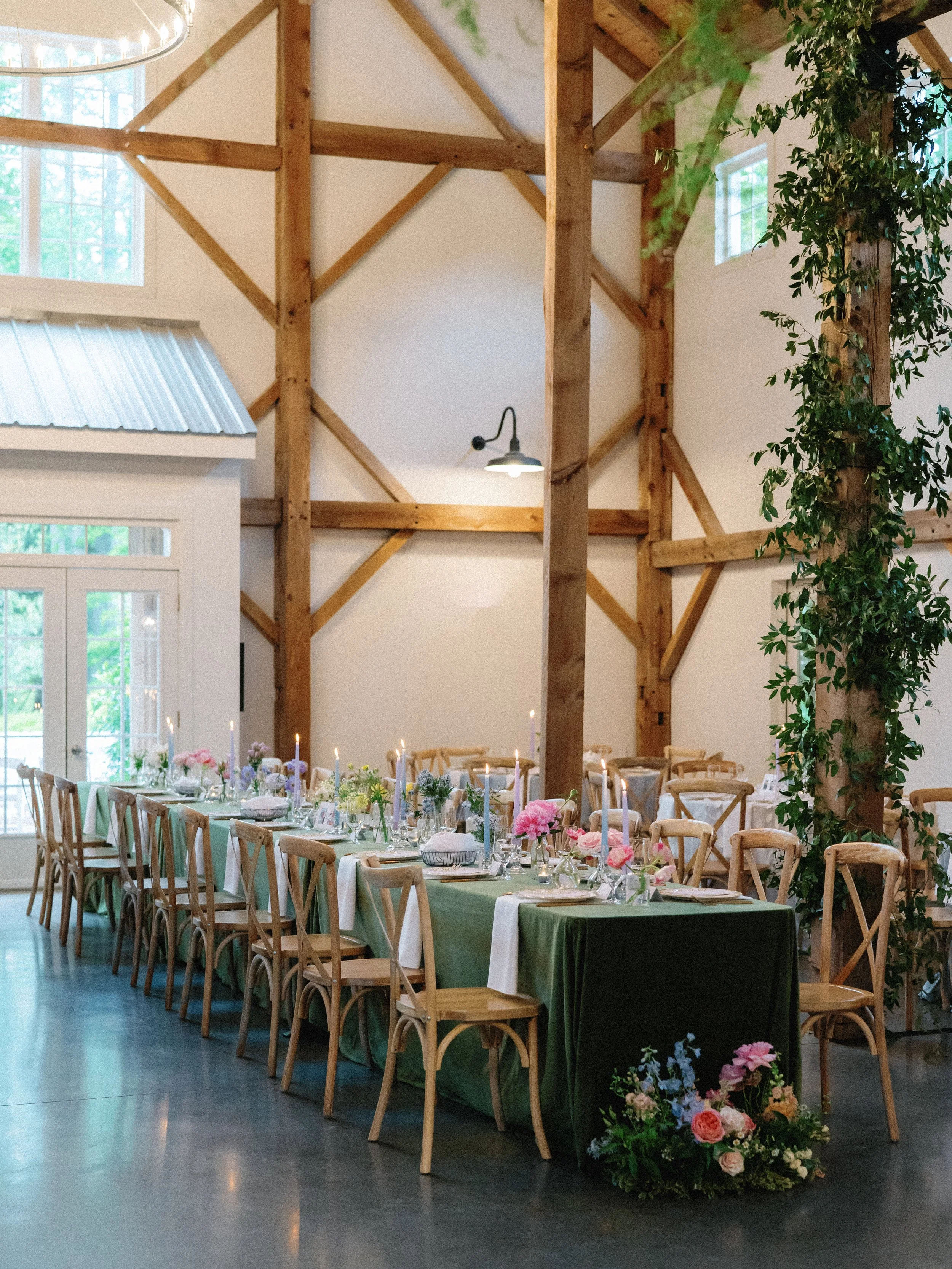 Long banquet table set up for a celebration in a rustic barn with wooden beams, decor includes flowers, candles, and tableware, with a large floral arrangement at one end of the table.