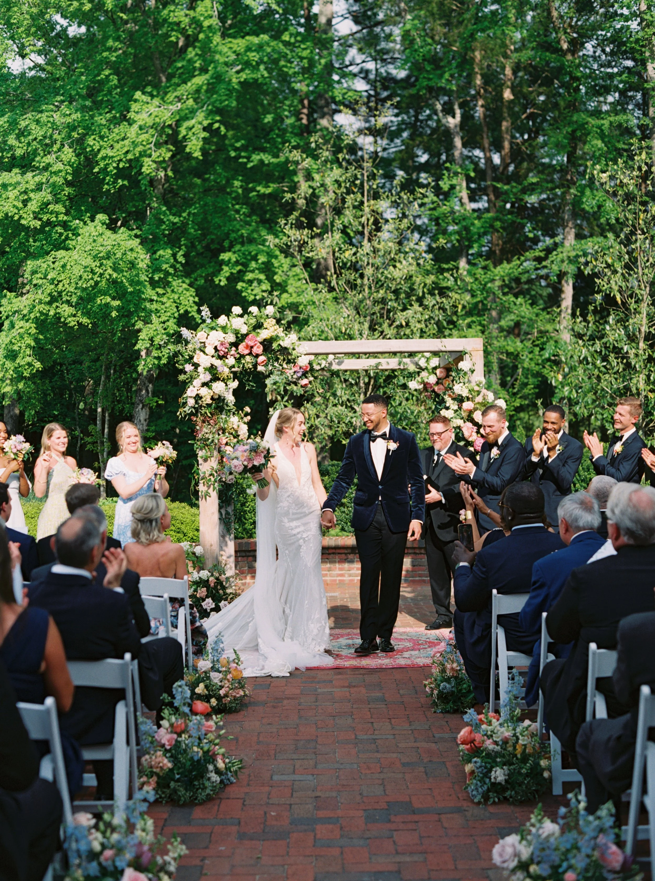 A wedding ceremony outdoors with the bride and groom holding hands, surrounded by wedding guests, under a floral arch, with lush green trees in the background.