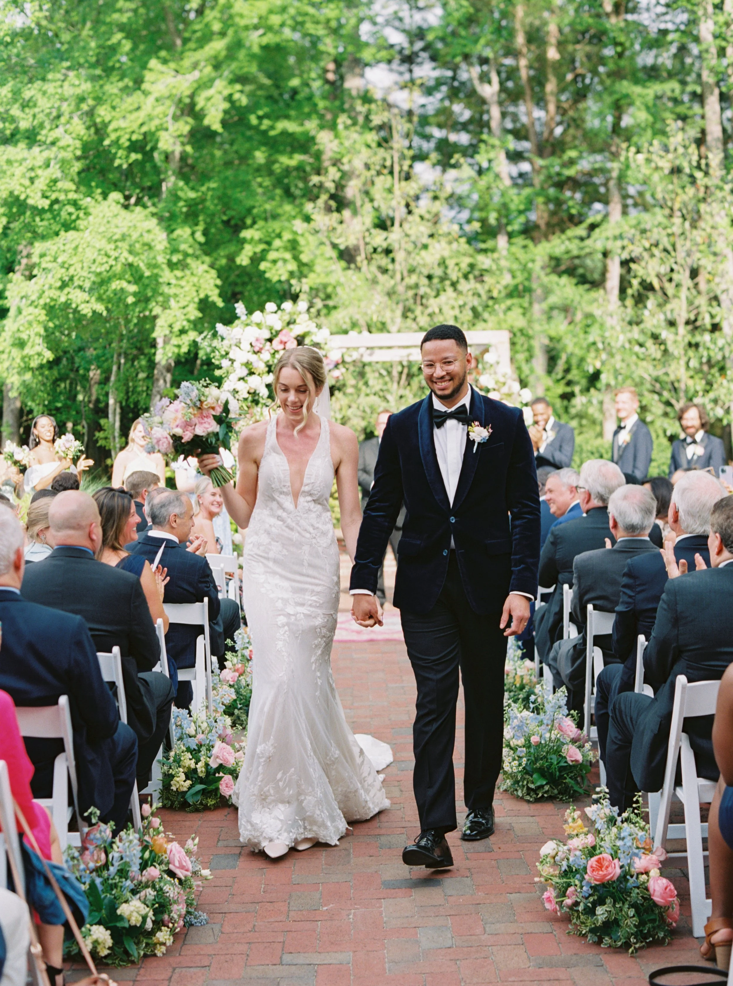 A newlywed couple walking down the aisle at their outdoor wedding, surrounded by family and friends, with floral arrangements and greenery in the background.