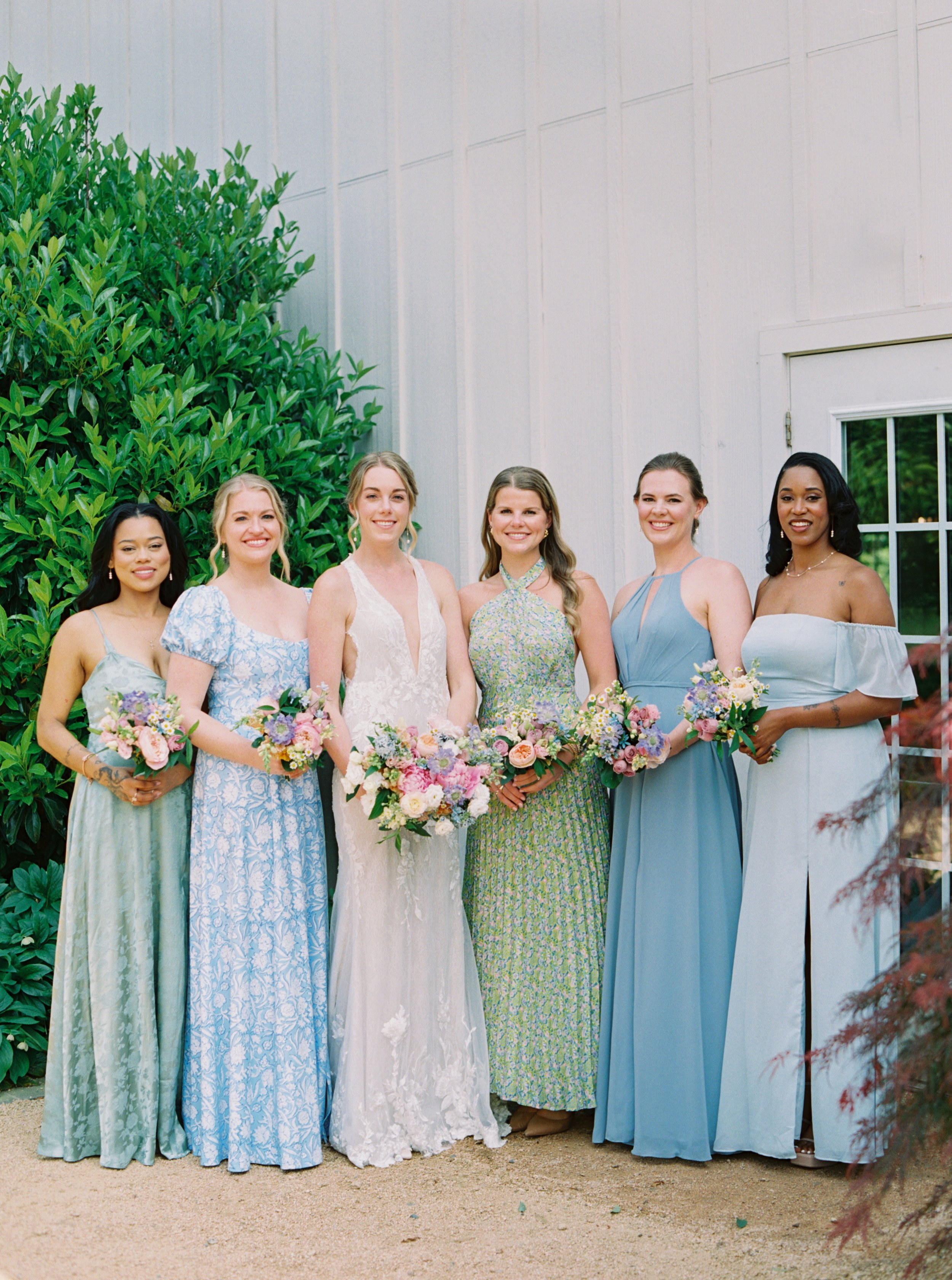 Group of six women dressed in colorful dresses holding bouquets, standing outdoors in front of a light wall and green foliage.