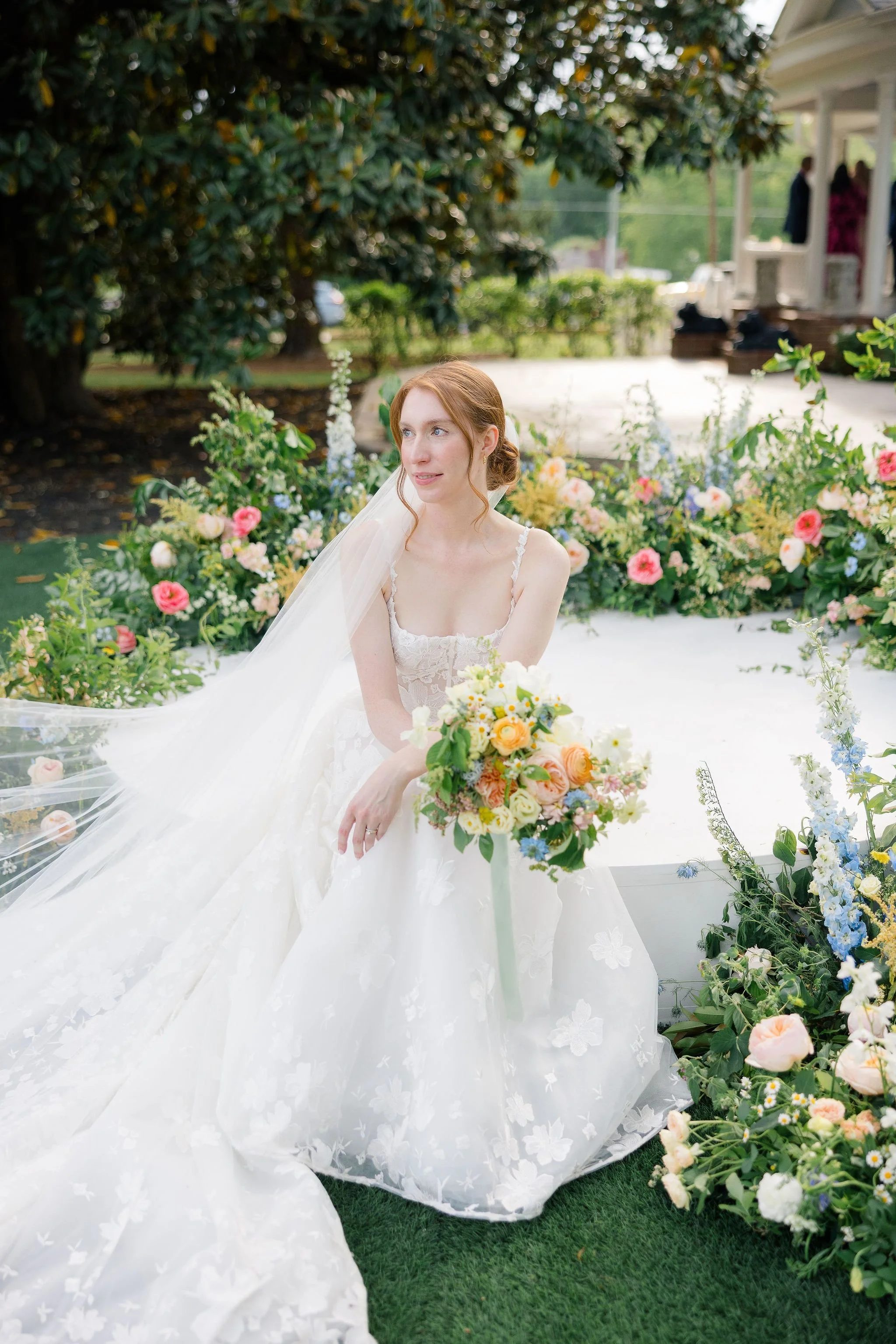 A bride in a white wedding dress sitting outdoors among colorful flowers with trees in the background.