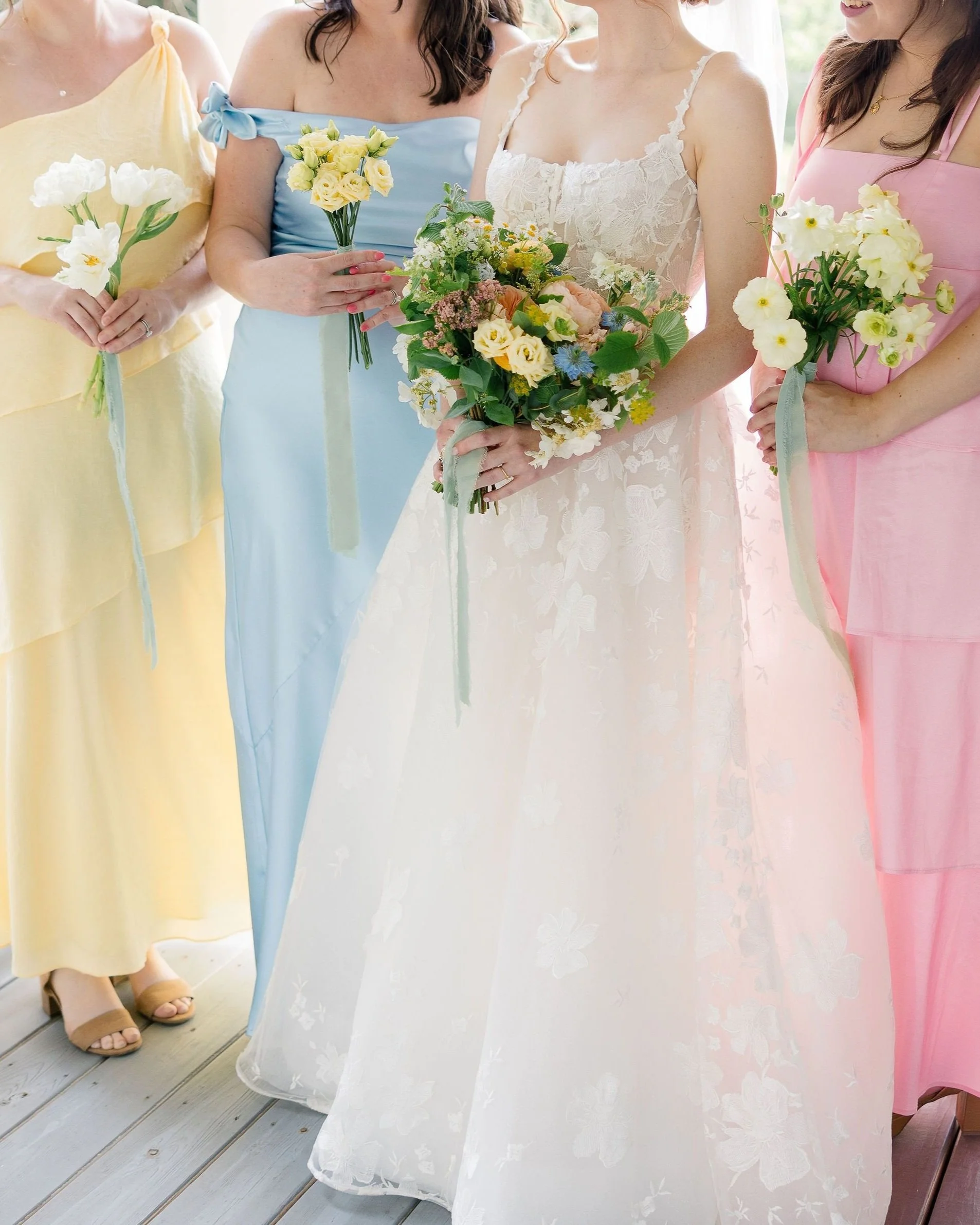Close-up of women wearing pastel-colored dresses, holding bouquets of flowers at a celebration or wedding. The focus is on the dresses and flowers, with the lower part of their bodies visible.