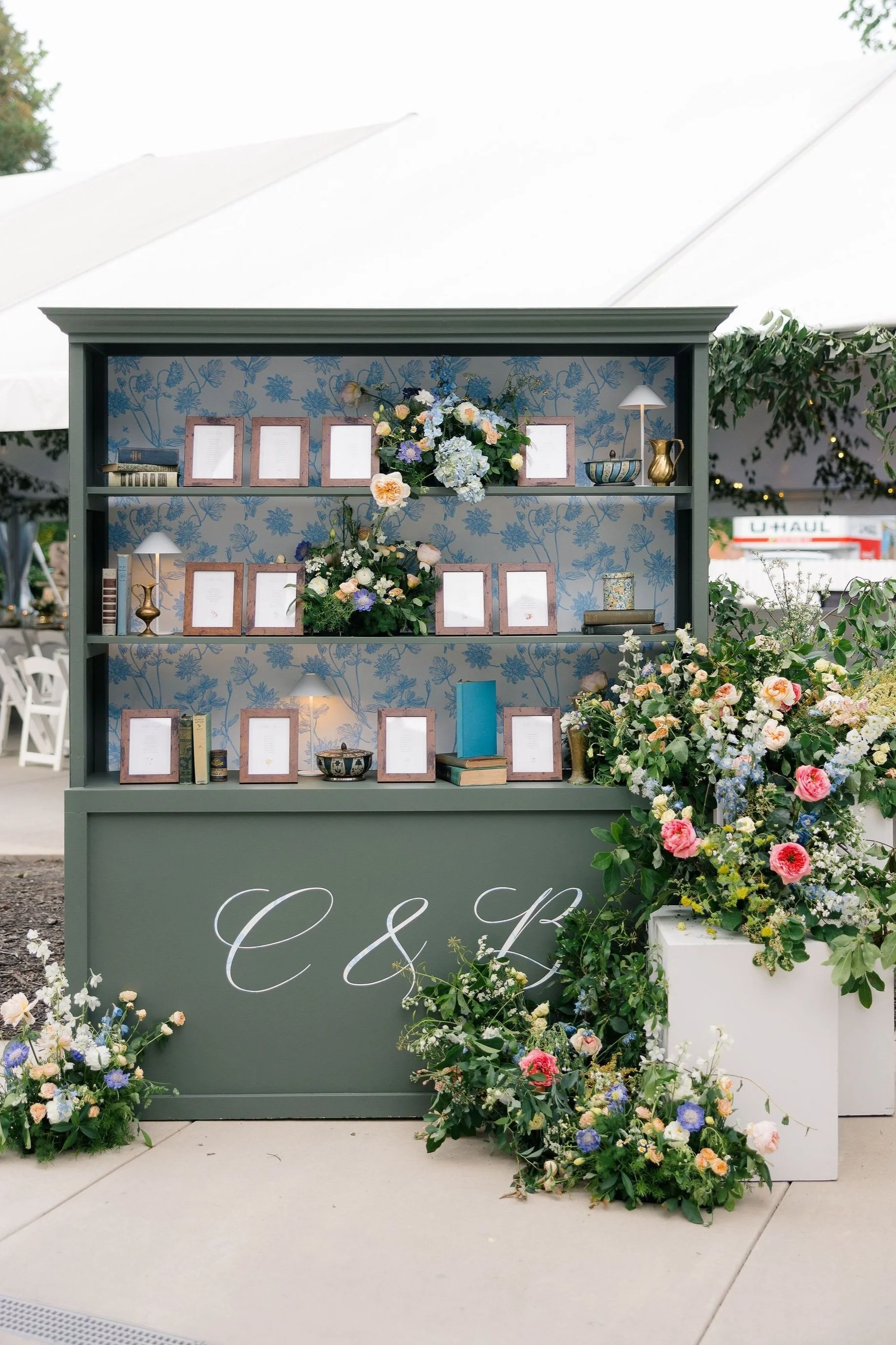 A decorative display shelf with blue floral wallpaper in the background, holding framed photos, books, flower arrangements, and small lamps, surrounded by lush flowers and greenery in an outdoor event setting.