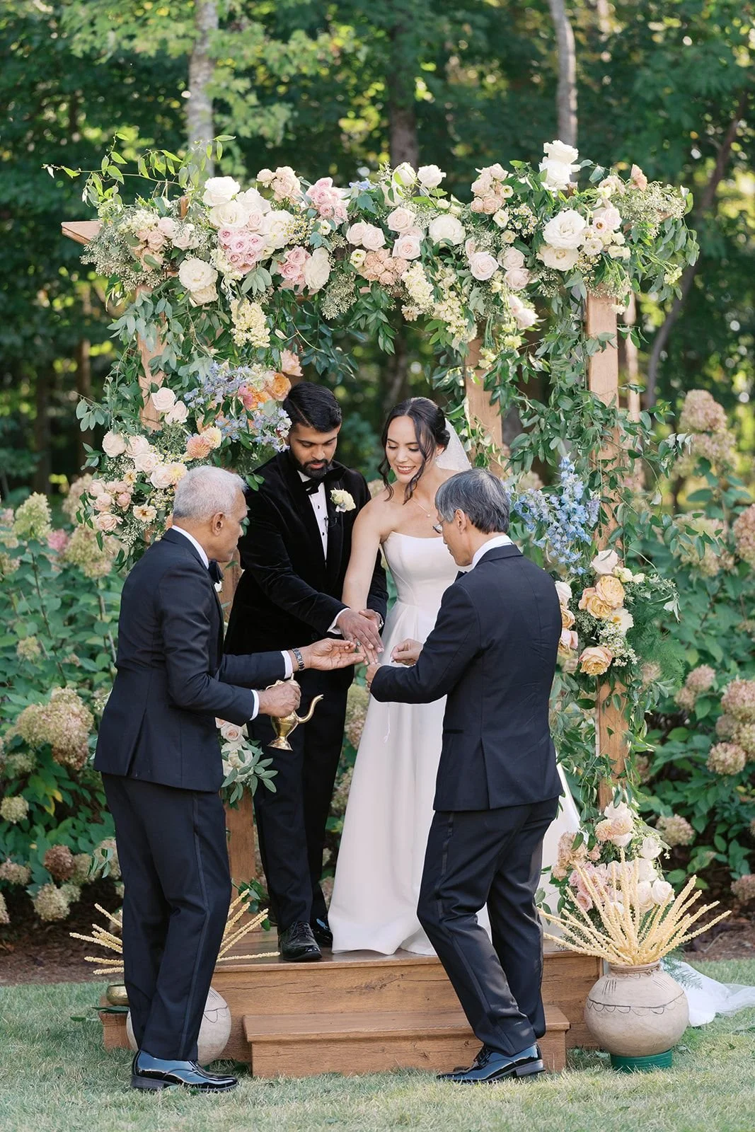 A wedding ceremony outdoors with a couple standing under a floral arch, exchanging rings, attended by two men in suits.