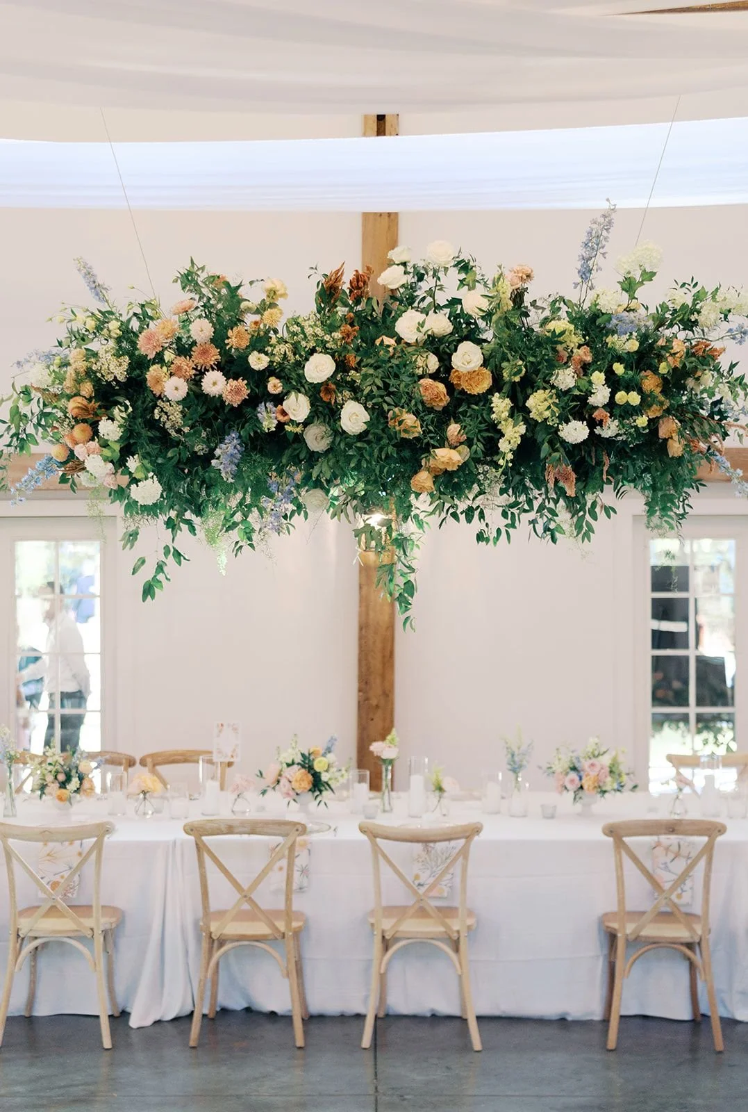 Indoor wedding reception with a long table decorated with floral arrangements and a large floral chandelier hanging above.
