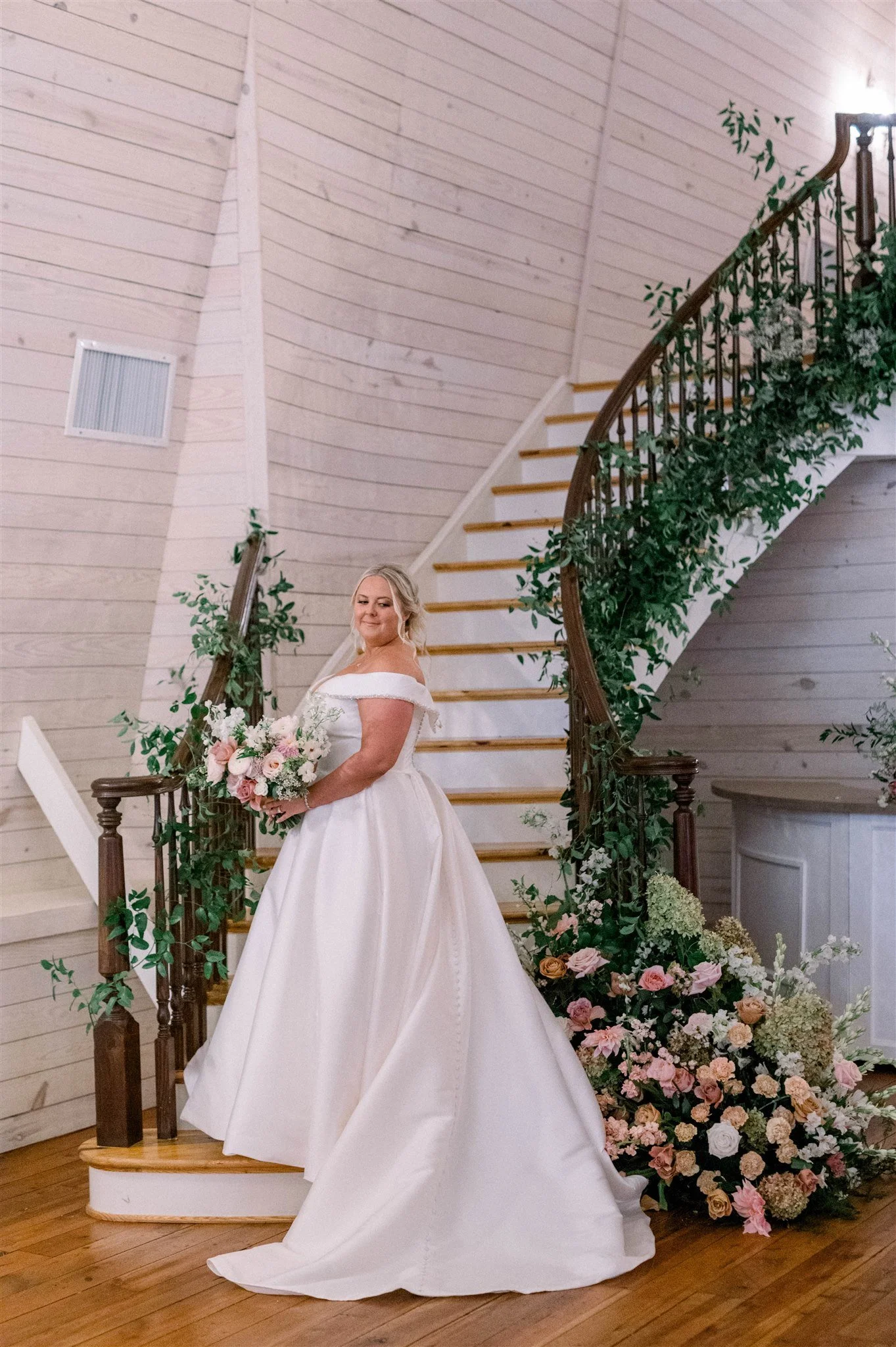 A bride in a white wedding dress holding a bouquet of flowers on a wooden staircase decorated with greenery and flowers.
