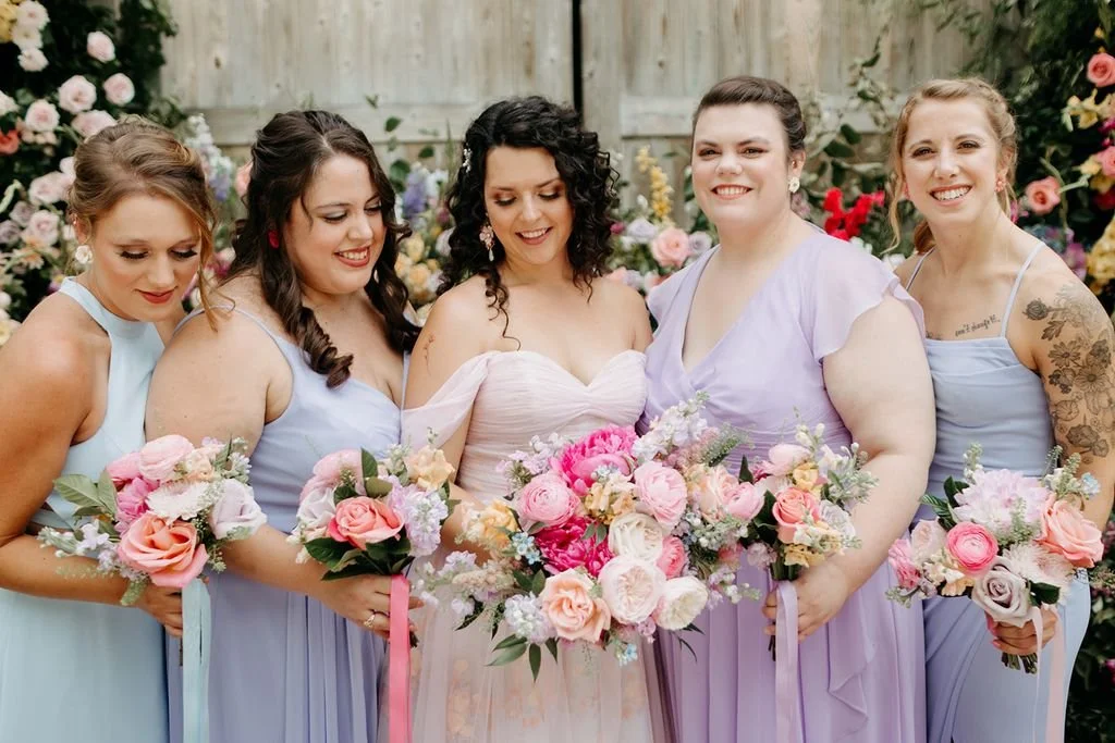 Group of women at a wedding, four bridesmaids and a bride, holding bouquets of pink, white, and purple flowers, standing outdoors in front of a wooden fence with floral decorations.