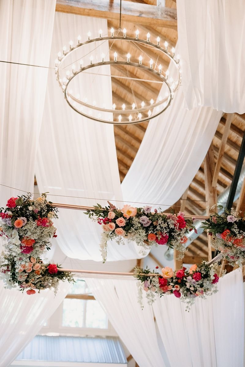 Decorated interior with a large circular chandelier and floral arrangements hanging from copper rods, white drapery, and wooden ceiling beams.
