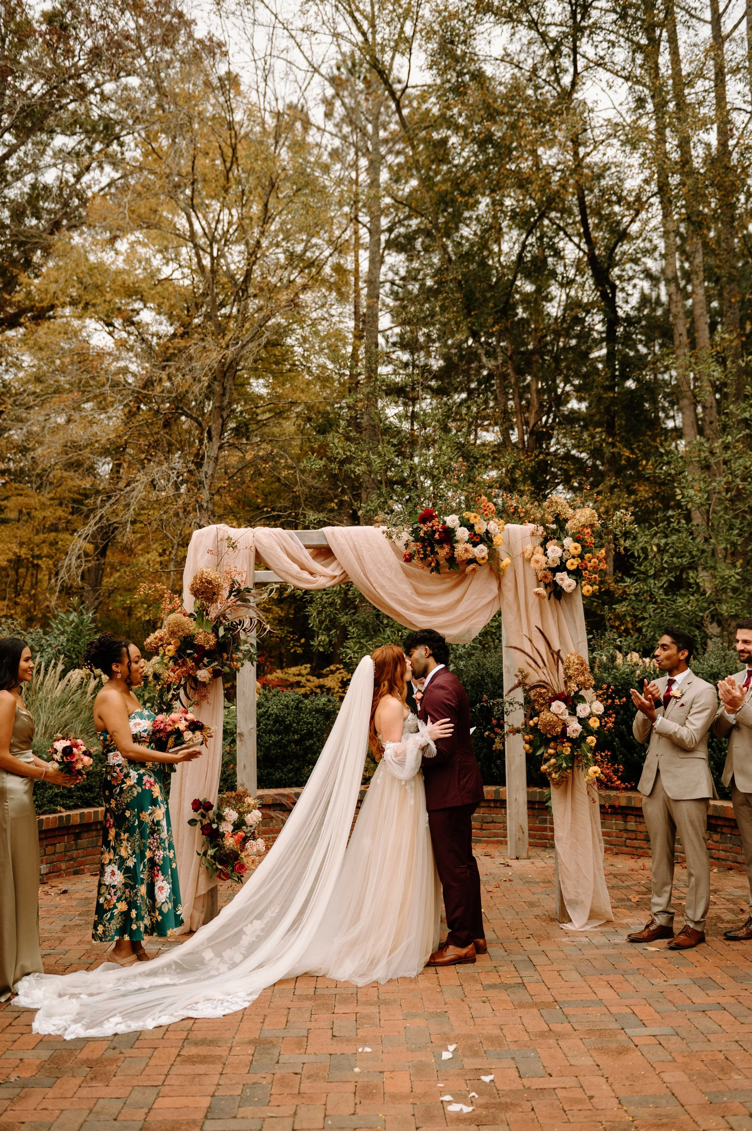 Couple sharing their first kiss during an outdoor wedding ceremony beneath a floral arch at The Barn of Chapel Hill
