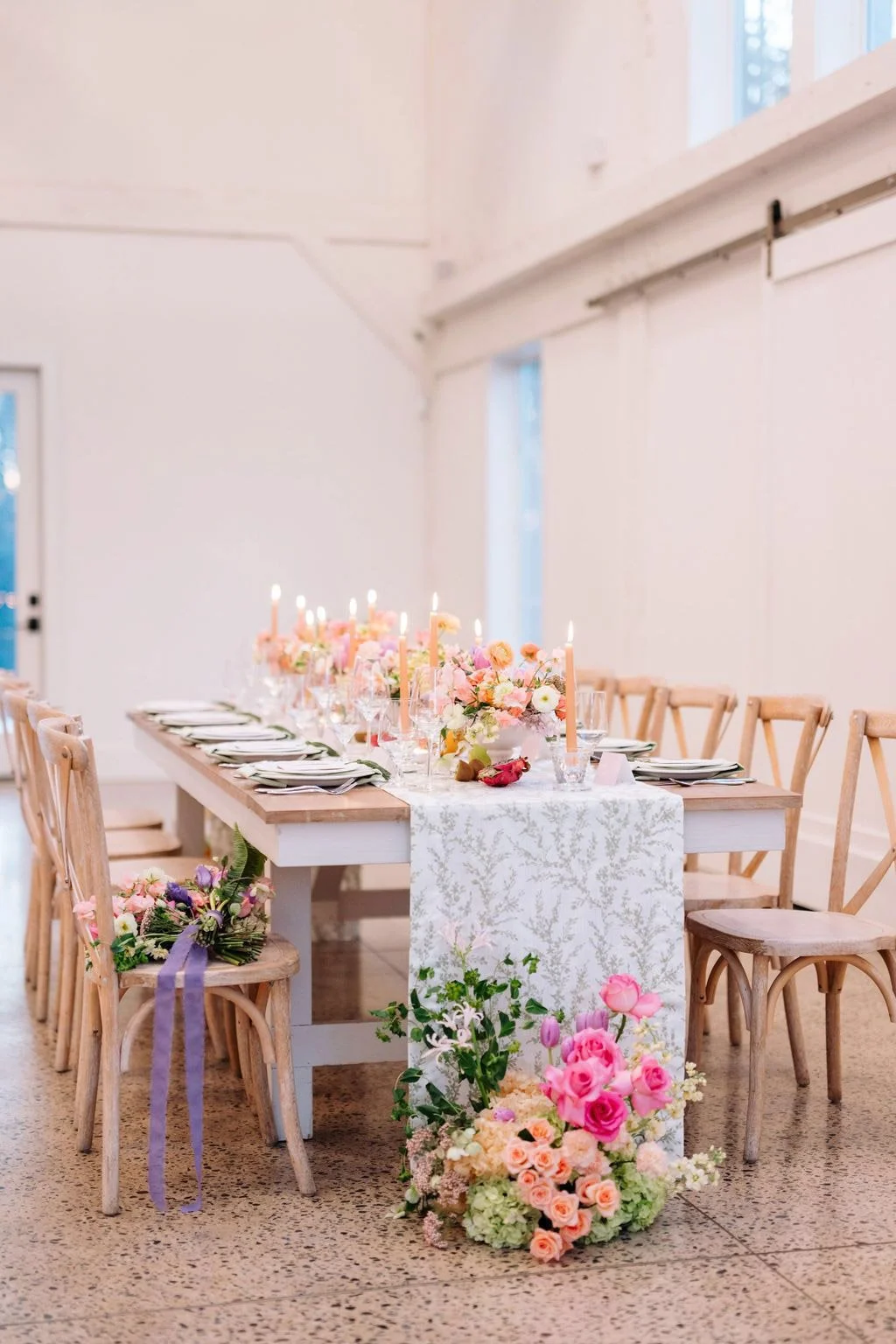 A beautifully set dining table decorated with pink and white flowers, candles, and place settings in a bright, minimalist room.