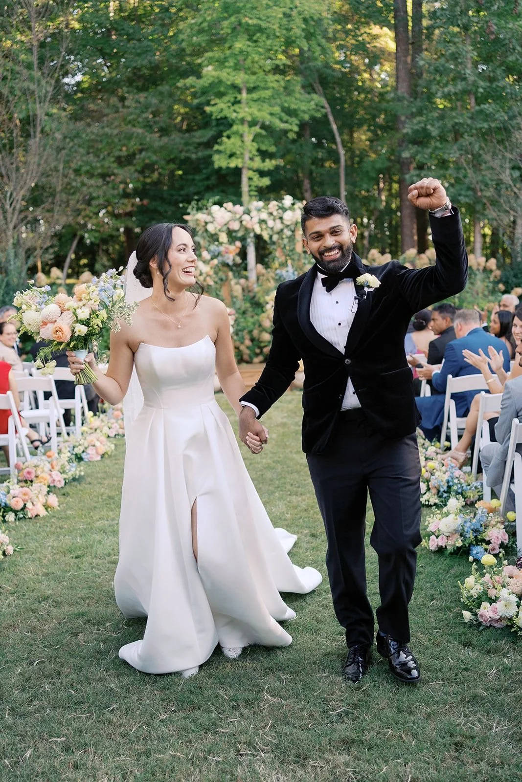 A newlywed couple holding hands and smiling, the bride holding a bouquet of flowers, walking down the aisle outdoors with guests seated on either side, in a garden setting surrounded by trees and floral decorations.