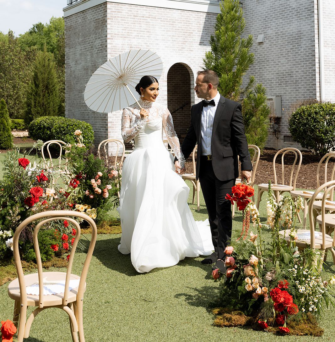 A bride and groom hold hands outdoors on a wedding day, with the bride holding a white parasol, surrounded by floral arrangements and empty chairs.