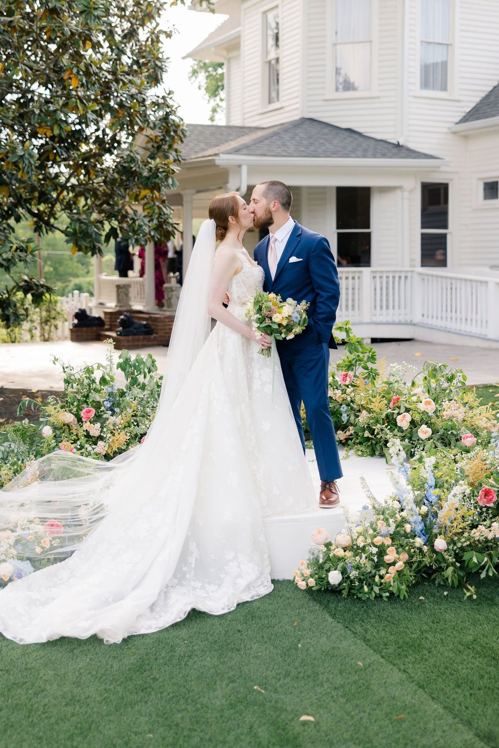 A bride and groom share a kiss outdoors during their wedding ceremony standing on a decorated platform with colorful flowers. The bride wears a white wedding gown with a long train and veil, holding a bouquet, while the groom is dressed in a blue suit with a pink tie, in front of a white house with a porch.