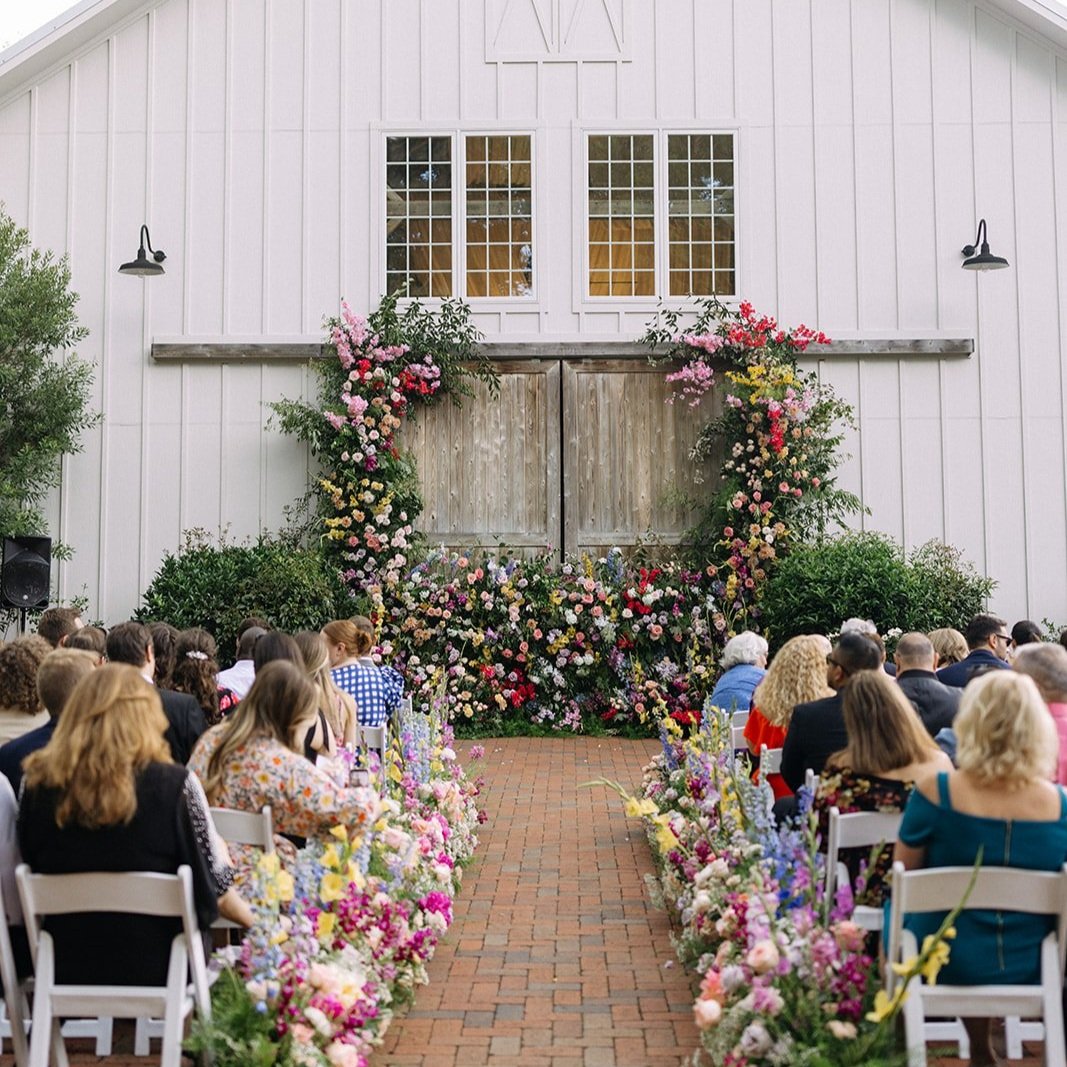 An outdoor wedding ceremony with chairs arranged along a brick aisle, decorated with colorful flowers, leading to a wooden door on a white building with floral arrangements around it, and guests seated facing the setup.