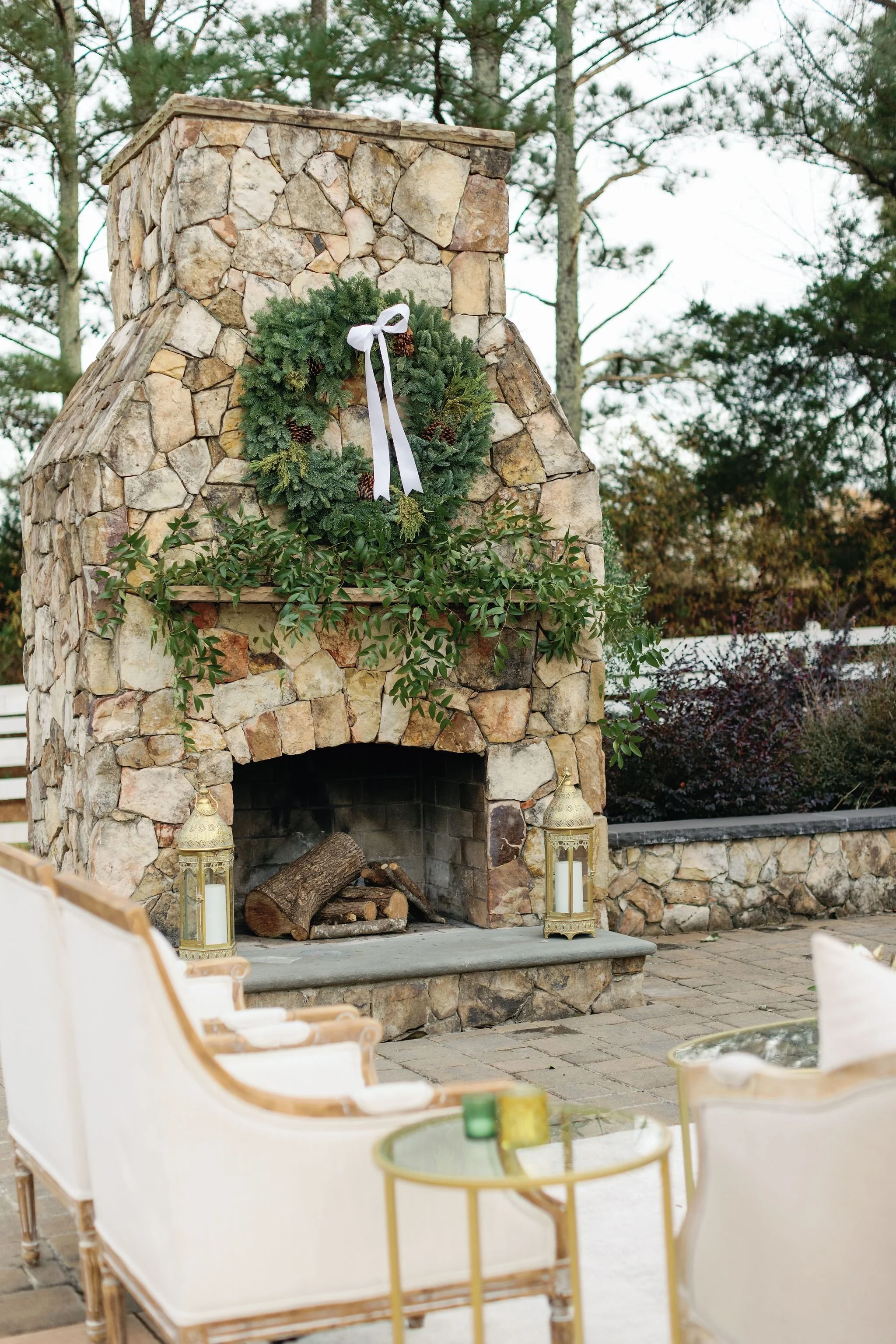 An outdoor stone fireplace decorated with a green wreath with pinecones and a white bow, with lanterns on each side and white chairs facing towards it, in a backyard setting with trees in the background.