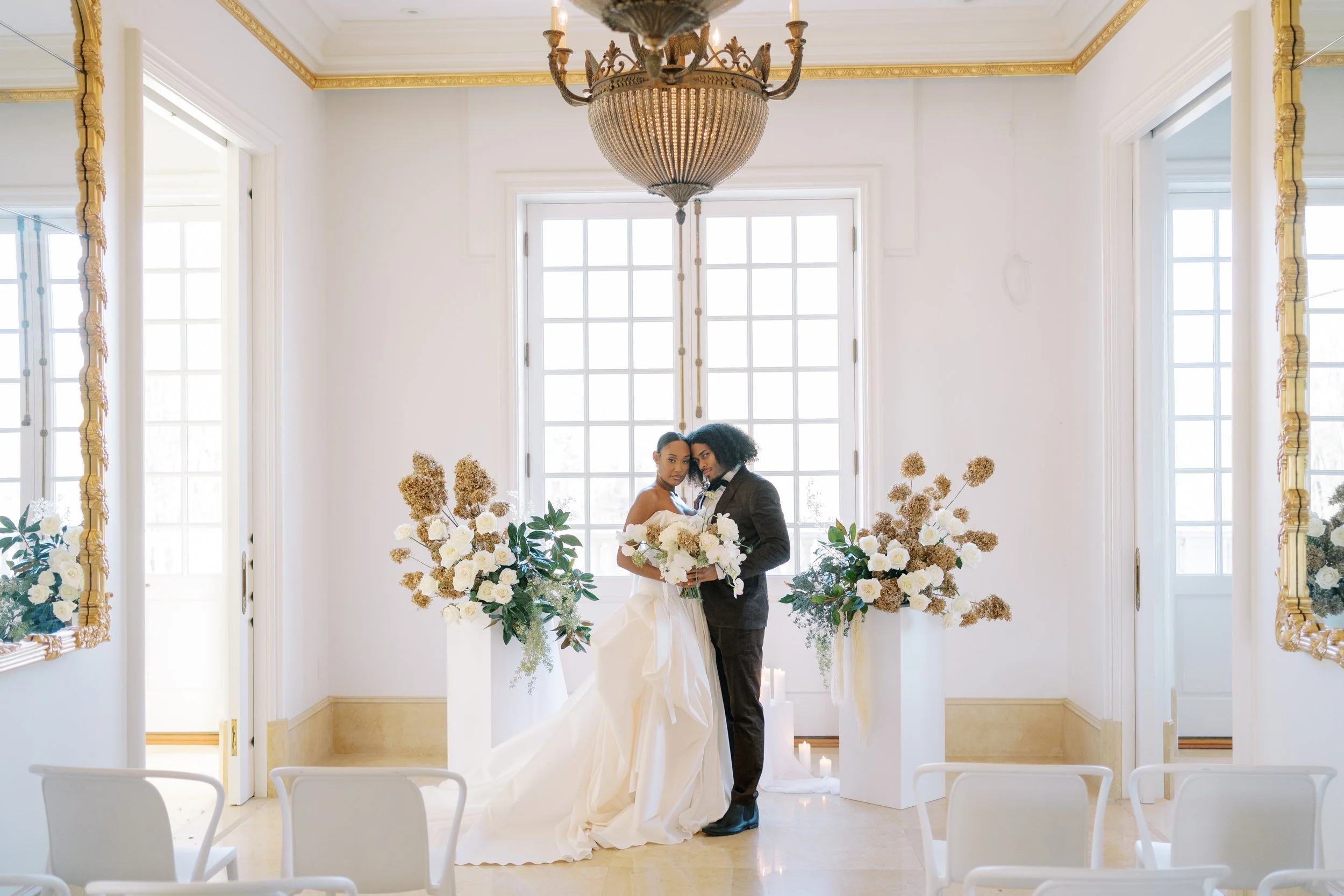A couple dressed in wedding attire standing close together with their foreheads touching in a bright, elegant room decorated with flowers and gold accents, with windows and a chandelier overhead.