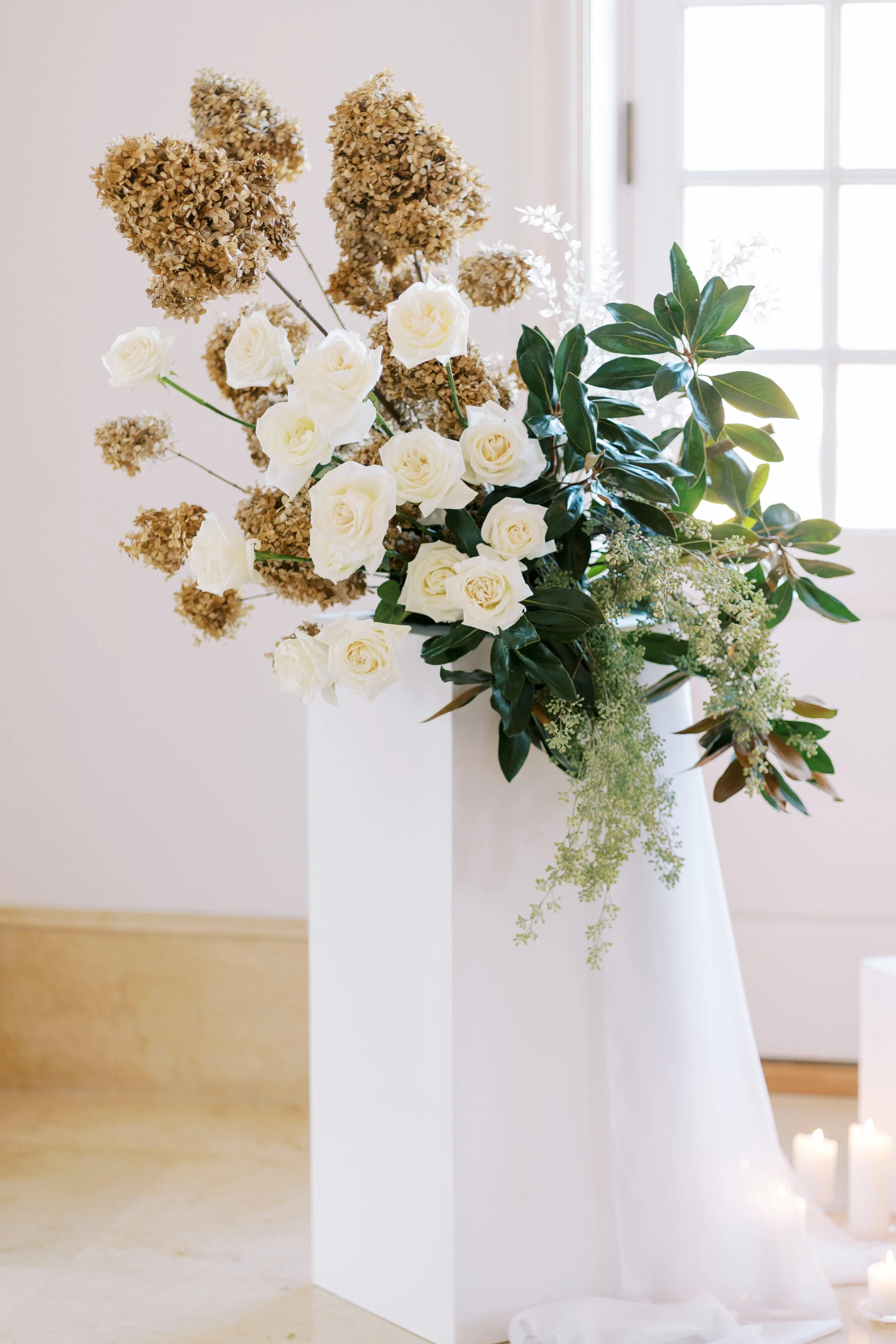 A floral arrangement with white roses, brown hydrangeas, and green leaves in a white vase, set against a bright window background.