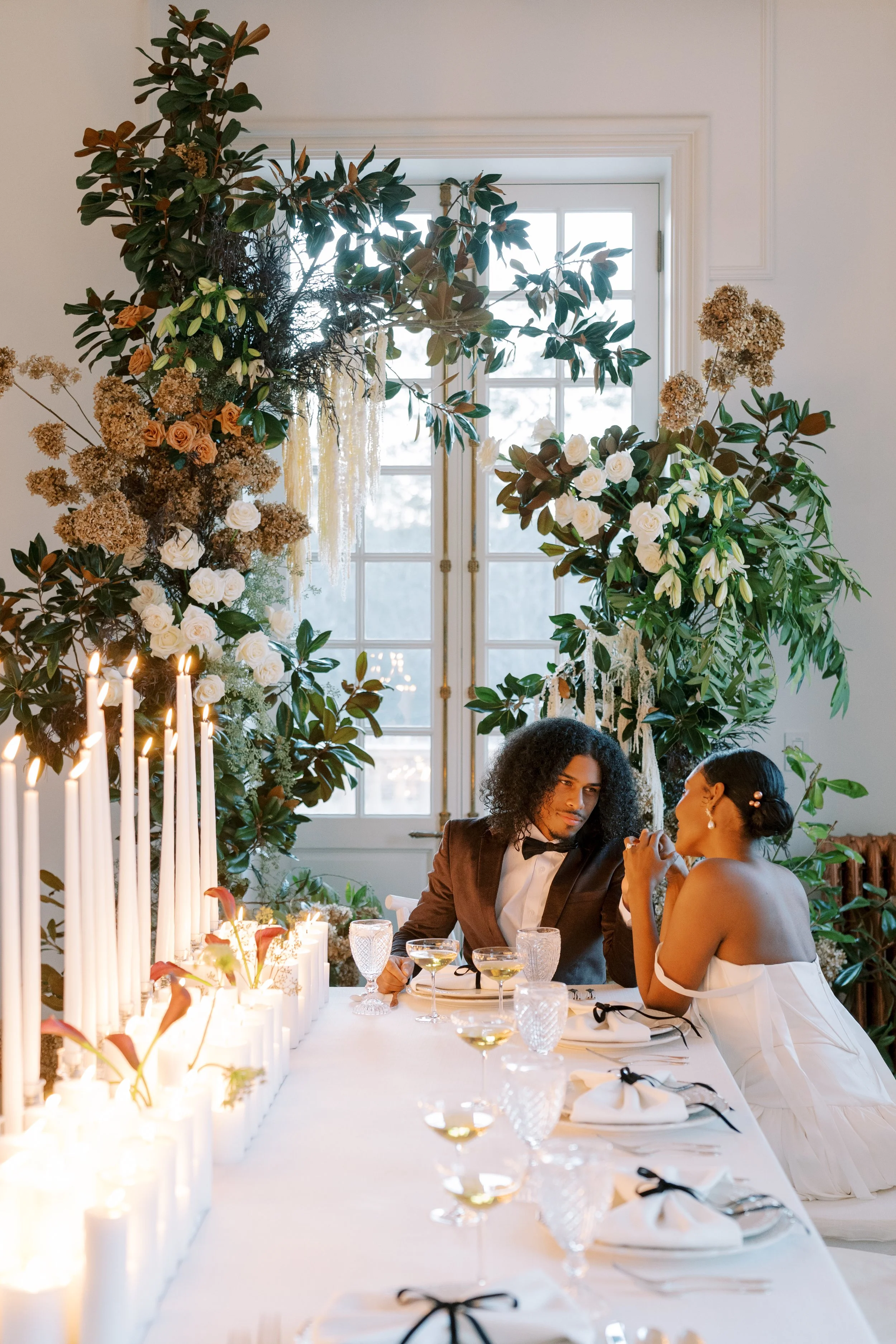 A couple is sitting and talking at a decorated dining table during a celebration or wedding reception, with tall candles and floral arrangements behind them and a window in the background.