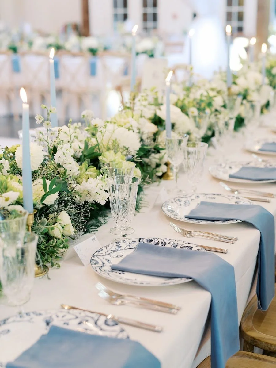 Elegant banquet table setup with white and blue color scheme, featuring floral centerpieces, candles, patterned plates, blue napkins, and crystal glassware.