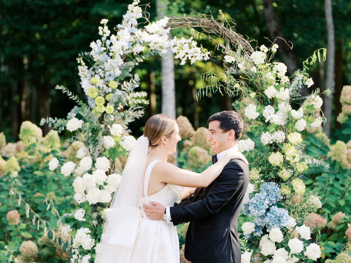 A bride and groom smiling and embracing each other at their outdoor wedding ceremony, surrounded by a floral arch with white, cream, and blue flowers.