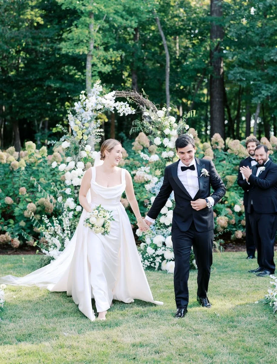 A newly married couple walking hand in hand outdoors at their wedding, surrounded by white and cream floral arrangements and greenery.