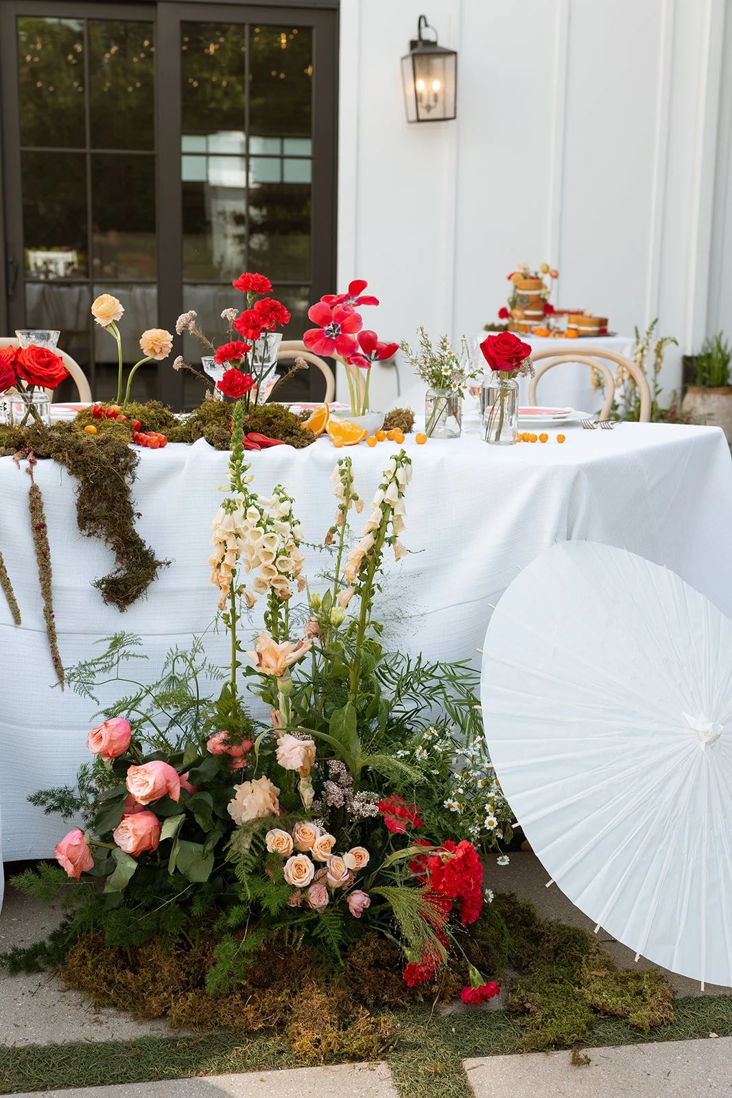 A decorated outdoor table with floral arrangements, candles, and a white tablecloth, set for a celebration or gathering, with a white wall, large window, and a lantern on the wall in the background.