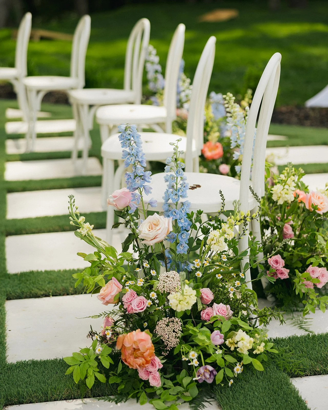 A row of white wooden chairs on a grassy outdoor setting, decorated with colorful flower arrangements including pink roses, blue delphiniums, and white daisies, suggesting a wedding or garden ceremony.