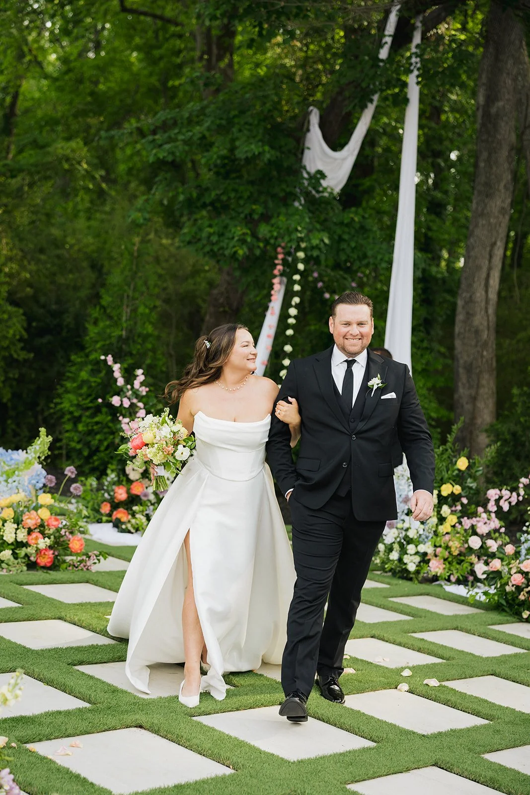A bride and groom walking outdoors after their wedding ceremony, surrounded by colorful flowers and greenery.