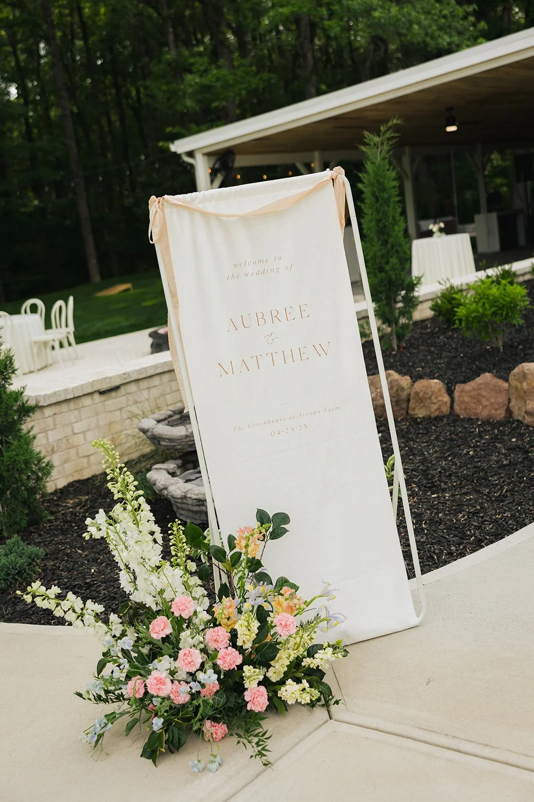 Wedding welcome sign with the names Aubree and Matthew, floral arrangement at the base, and outdoor venue in the background.