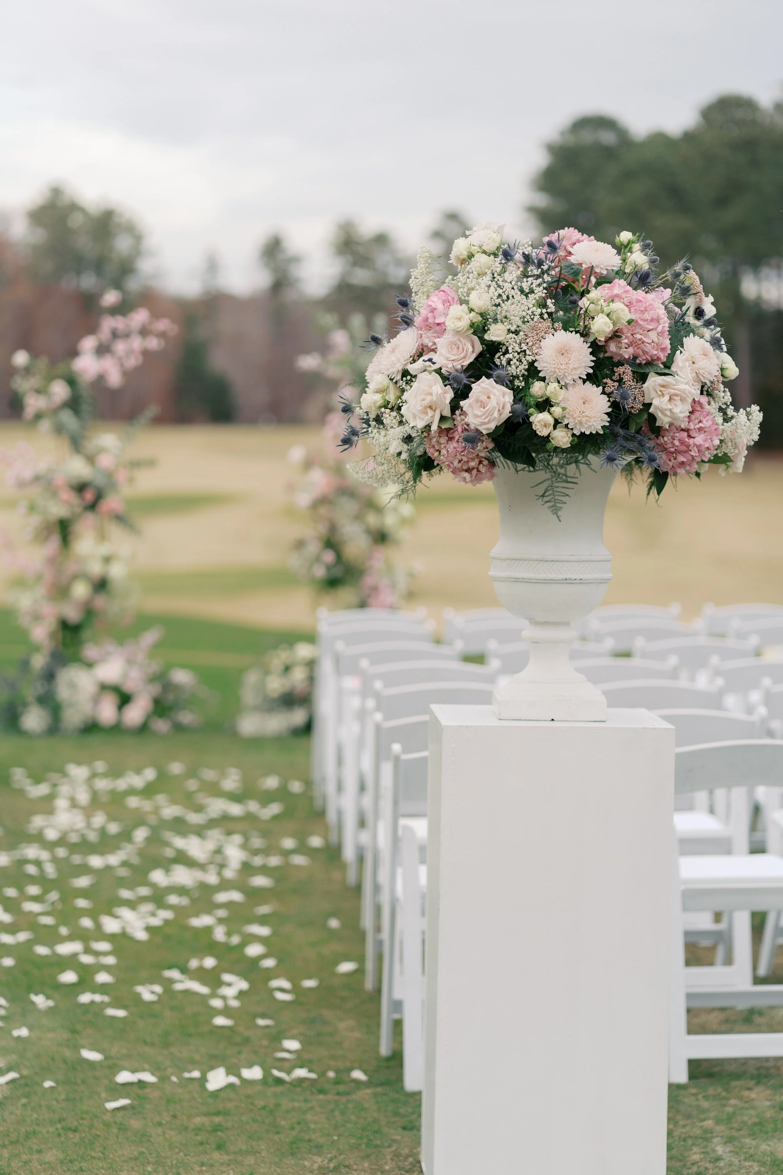 A floral arrangement in a white urn on a stand, with rows of white chairs and scattered flower petals on a grassy outdoor wedding ceremony setup.