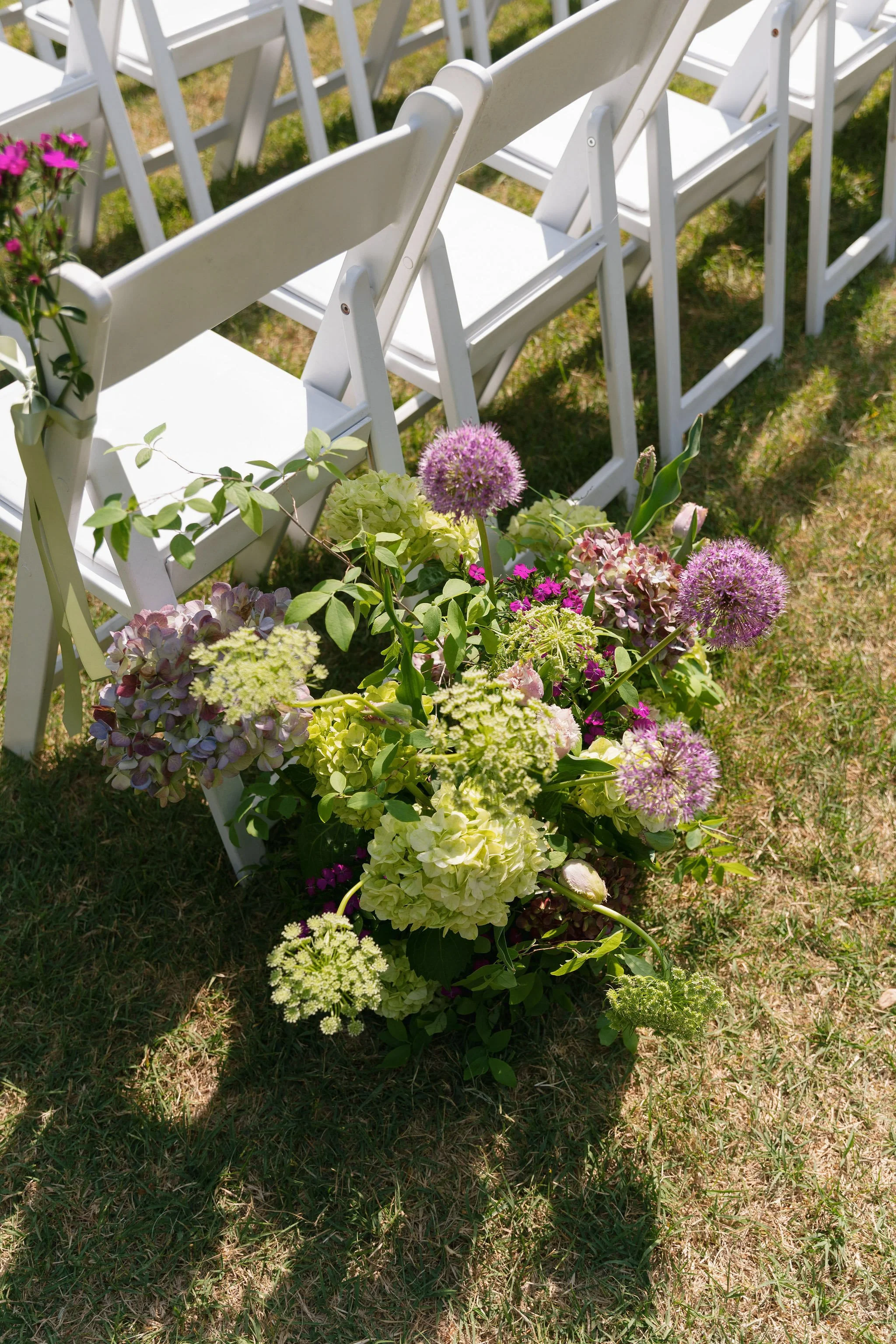  Meadow-style aisle arrangement of green hydrangea, purple allium and sweet william beside white ceremony chairs 