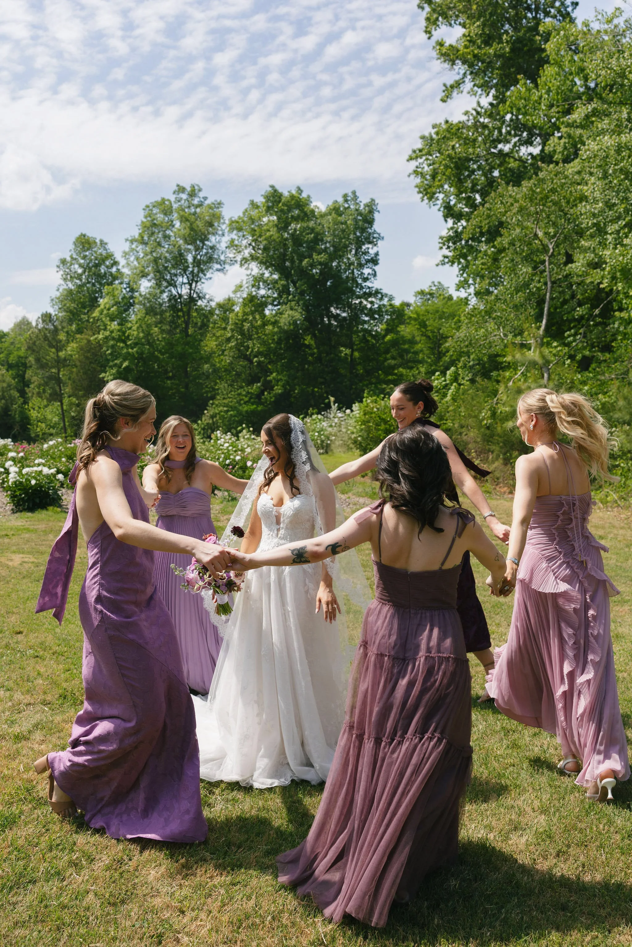  Bride laughing with her bridesmaids in shades of purple in a flower field at The Barn of Chapel Hill 