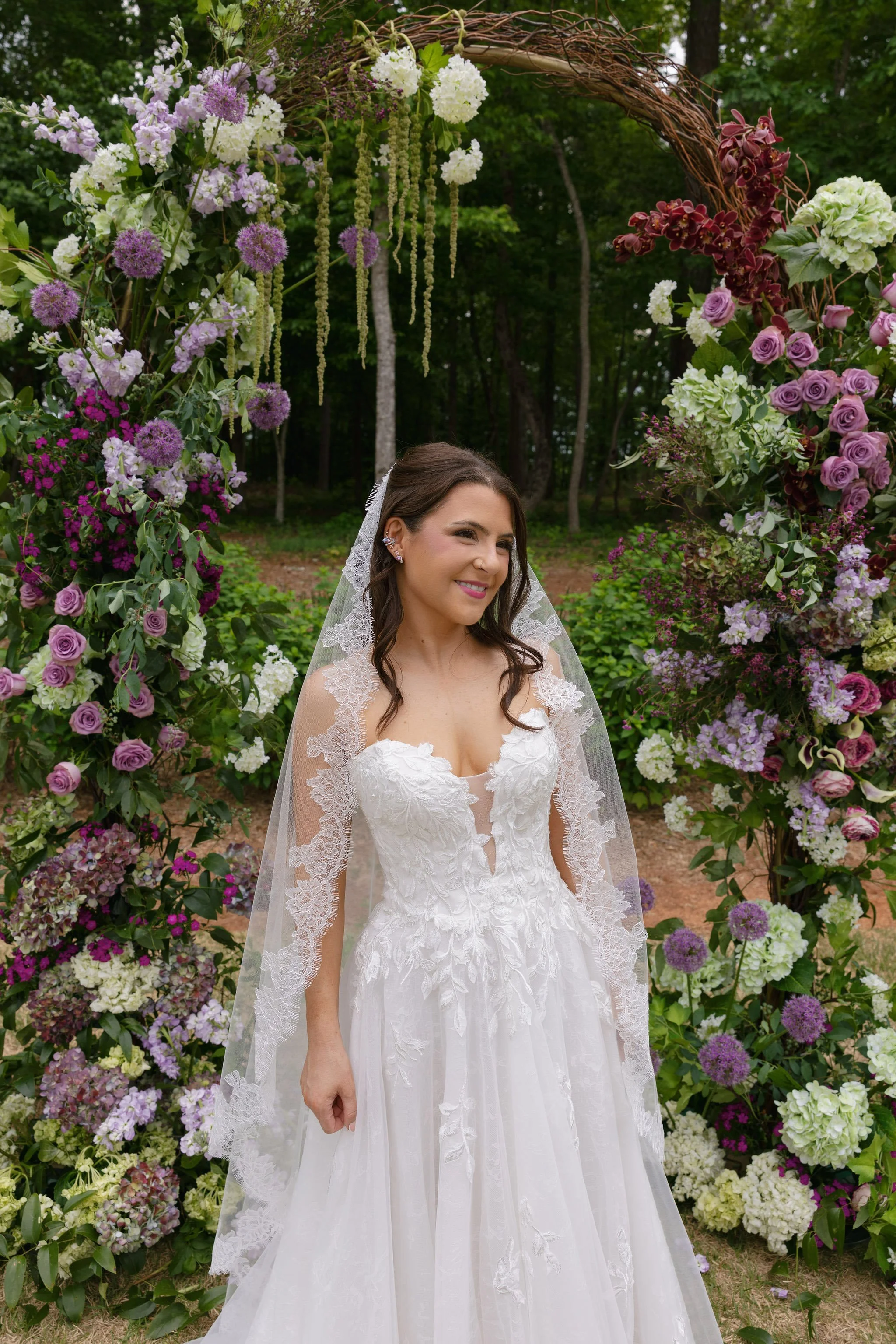  Bride in a lace A-line gown and lace-edged veil smiling beside a grapevine arch full of lavender roses, hydrangea, allium, and burgundy orchids at The Barn of Chapel Hill 