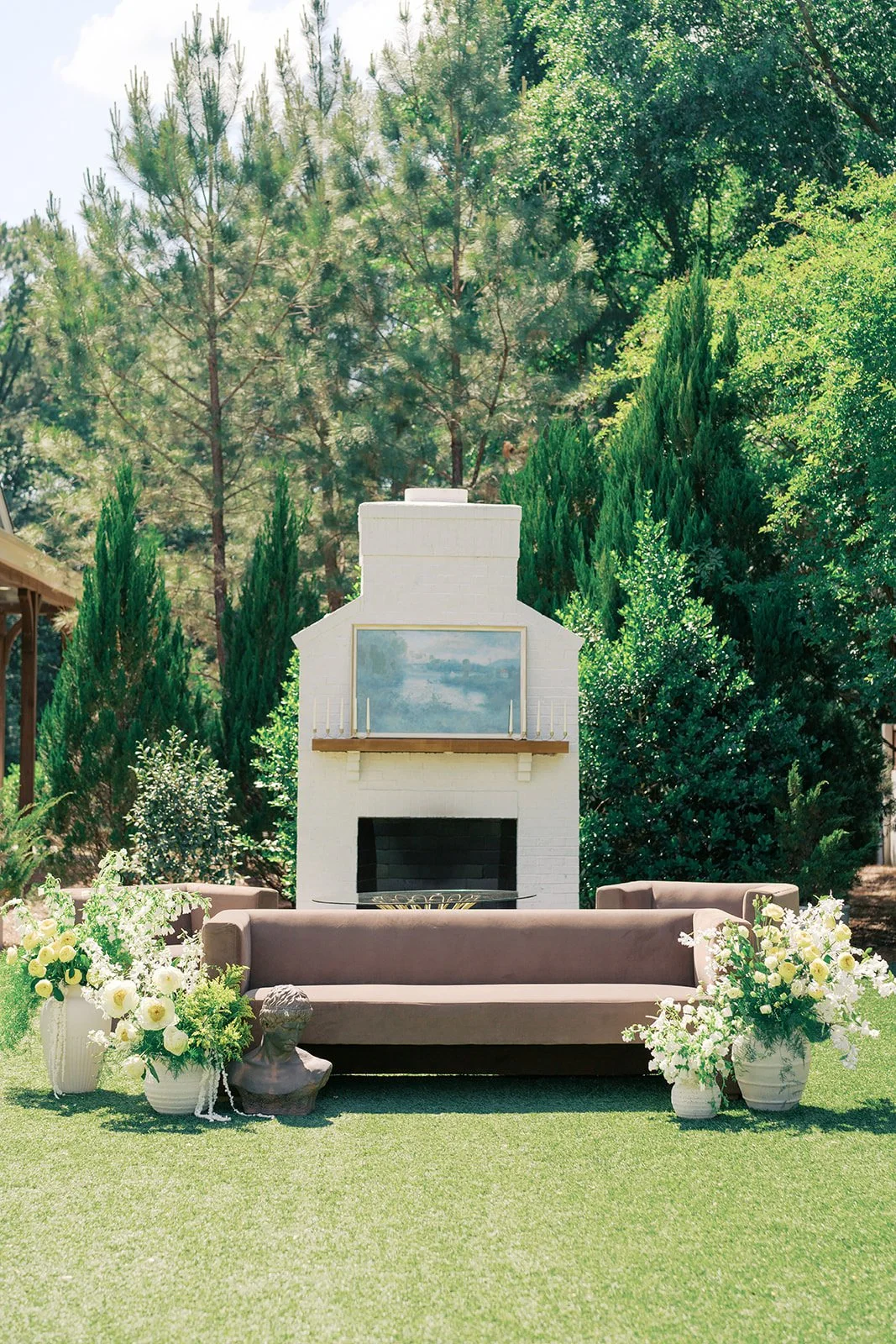  Outdoor fireplace lounge with a velvet sofa flanked by white garden roses and greenery on the lawn at The Bradford 