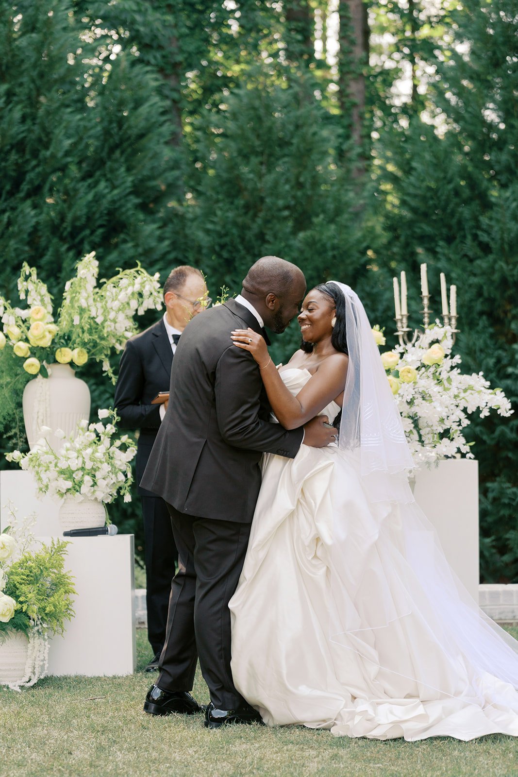  Bride and groom embracing at their ceremony framed by white floral urns and greenery on pedestals at The Bradford 