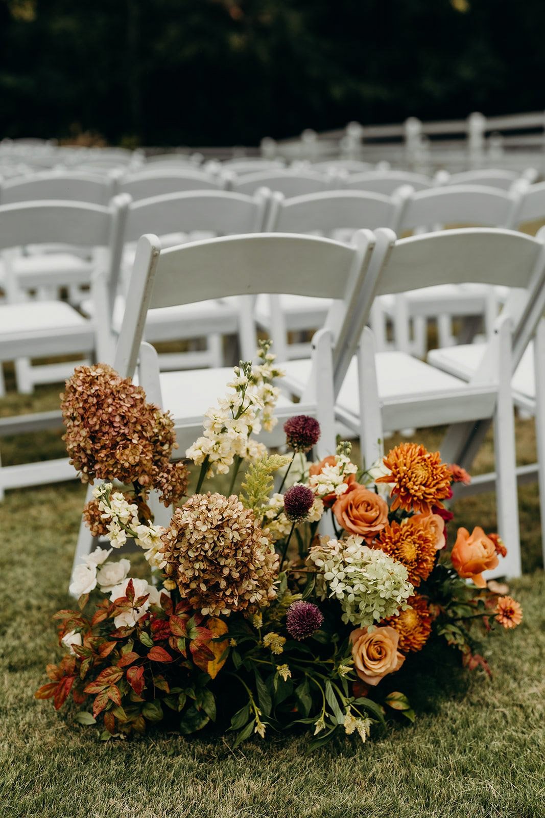 Fall ceremony aisle arrangement with dried limelight hydrangea, terracotta roses, orange chrysanthemums, and allium beside white chairs at The Wakefield Barn near Raleigh