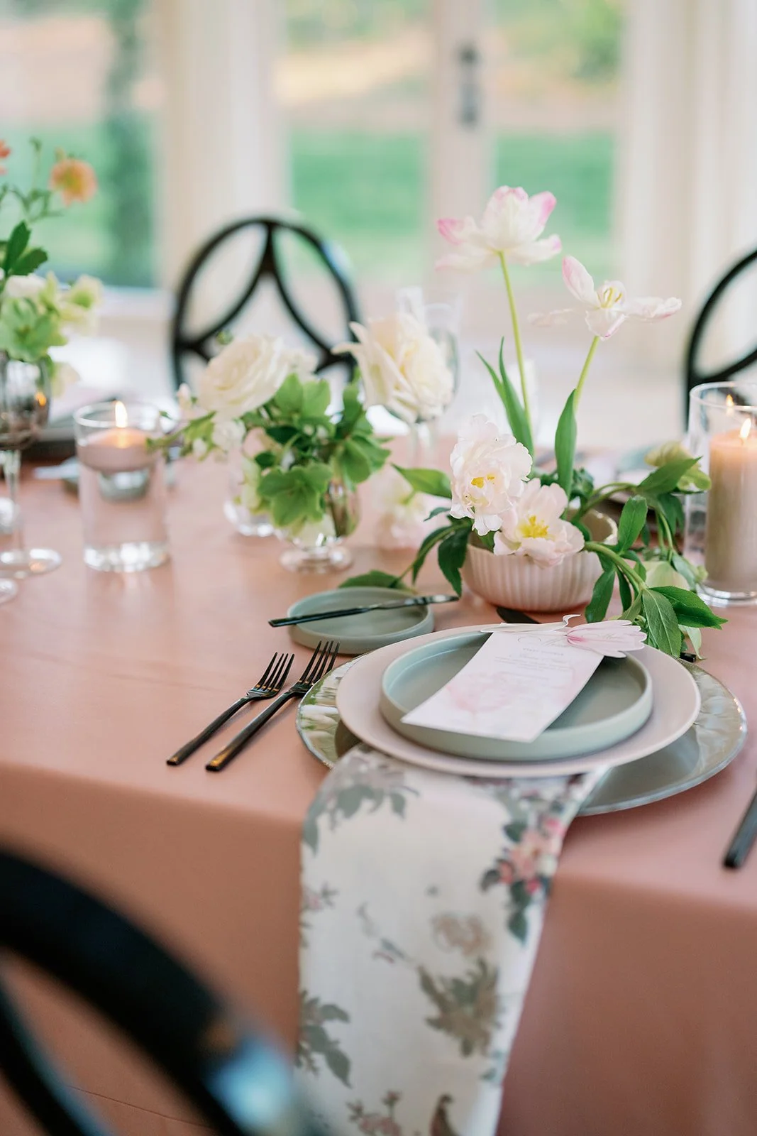 Elegant dining table with pastel pink tablecloth, floral centerpiece with white and pink flowers, candles in glass holders, and place settings with plates, forks, and a printed menu card.