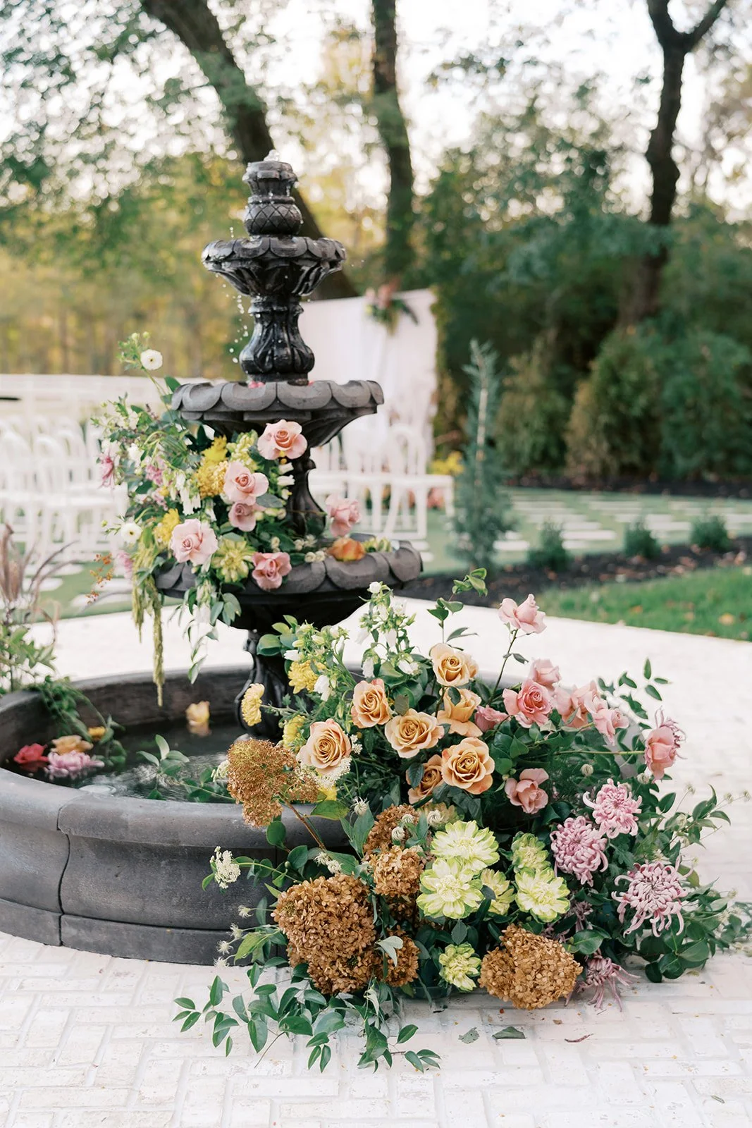 A black tiered garden fountain decorated with pink, yellow, and white flowers, surrounded by more flowers at its base, outdoors with trees and empty white chairs in the background.