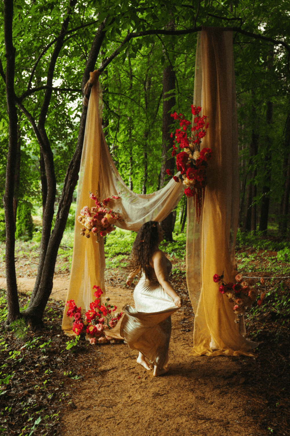 A woman with long, curly hair in a white dress dancing in a forest clearing, with a floral and fabric backdrop decorated with pink and red flowers, surrounded by lush green trees.