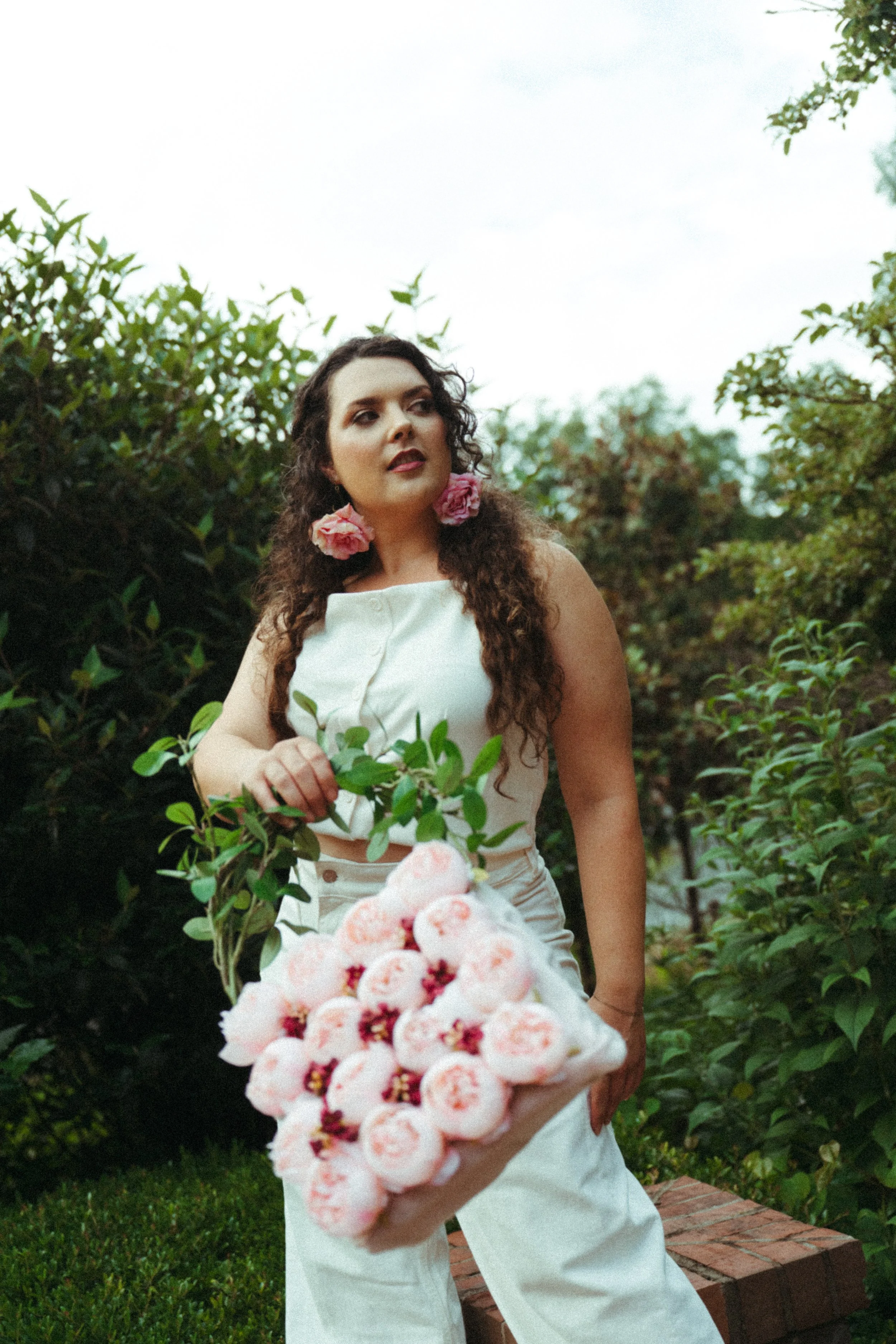 Woman with dark curly hair wearing large pink floral earrings, white sleeveless top, and white pants, standing outdoors among green bushes and trees, holding a bouquet of pink and white flowers.