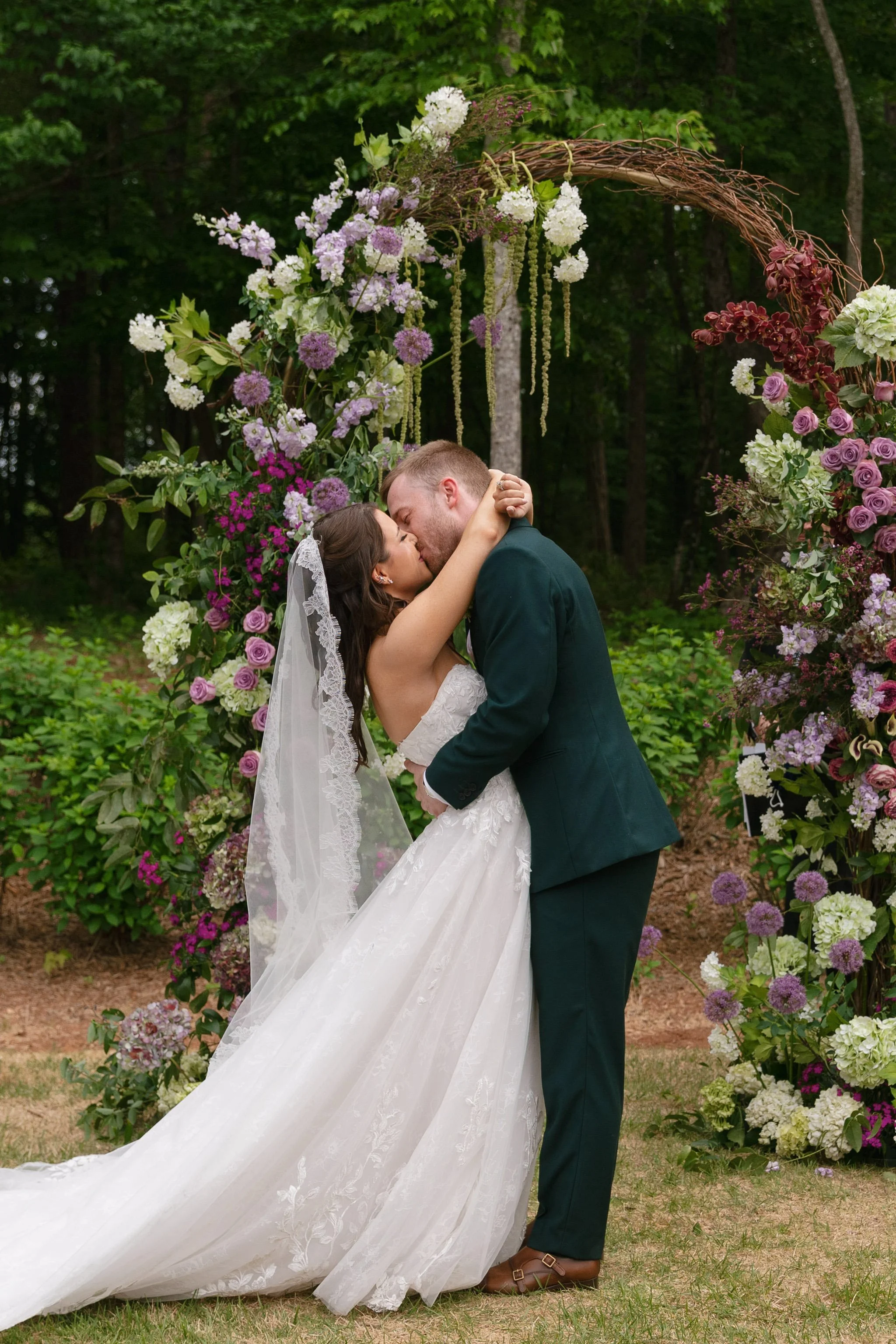 Newlyweds first kiss beside a grapevine floral arch in purple and green