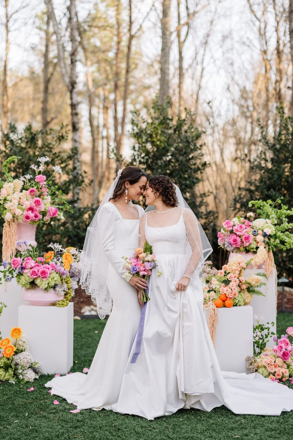 Two brides in white wedding dresses standing close and touching their foreheads together, holding bouquets of flowers, surrounded by colorful floral arrangements outdoors in a garden, with trees in the background.